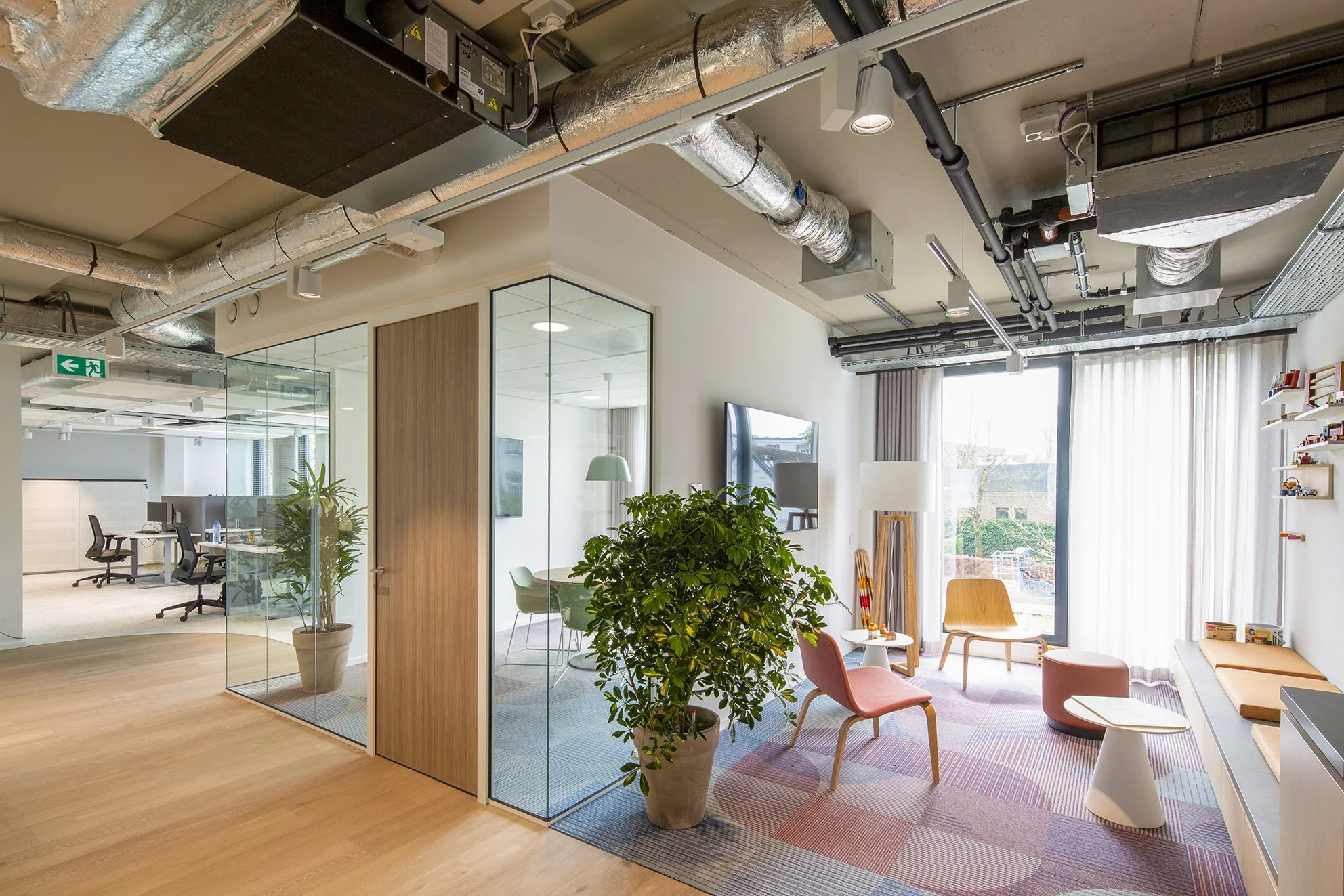 Lichte moderne kantoorruimte met grote ramen, planten, vergadertafel, en relaxruimte met stoelen en tafels, plafond met zichtbare ventilatiesystemen