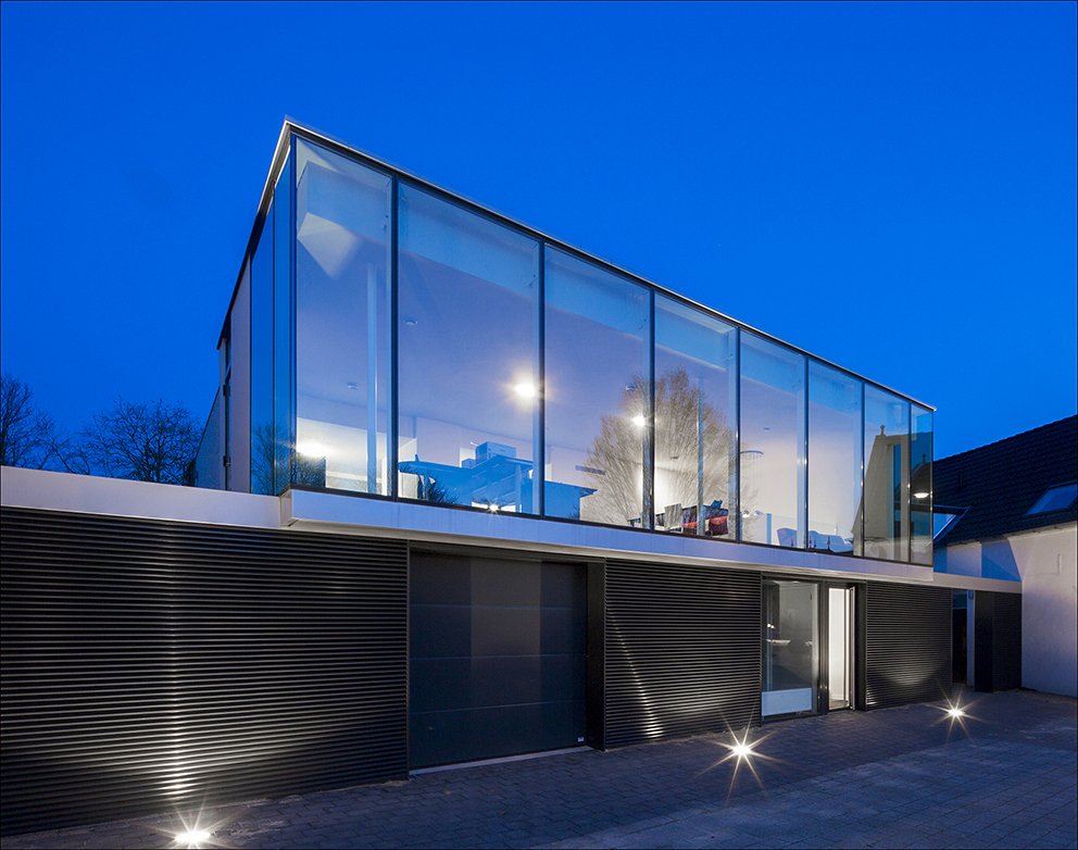 Moderne twee-verdiepings huis met grote glazen ramen, 's avonds verlicht, zwarte gevelbekleding en een glazen deur.