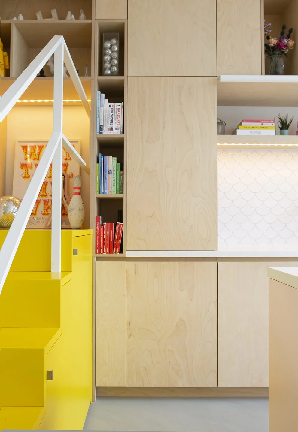 Een modern interieur met houten kasten, boeken, decoraties, bloemen, en een gele trap of kast.