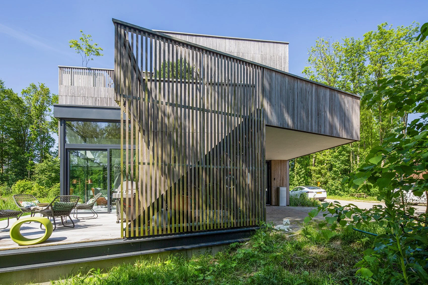 Moderne houten huis met grote ramen en buitenterras, omgeven door groen bos, met een auto geparkeerd onder een uitbouw.