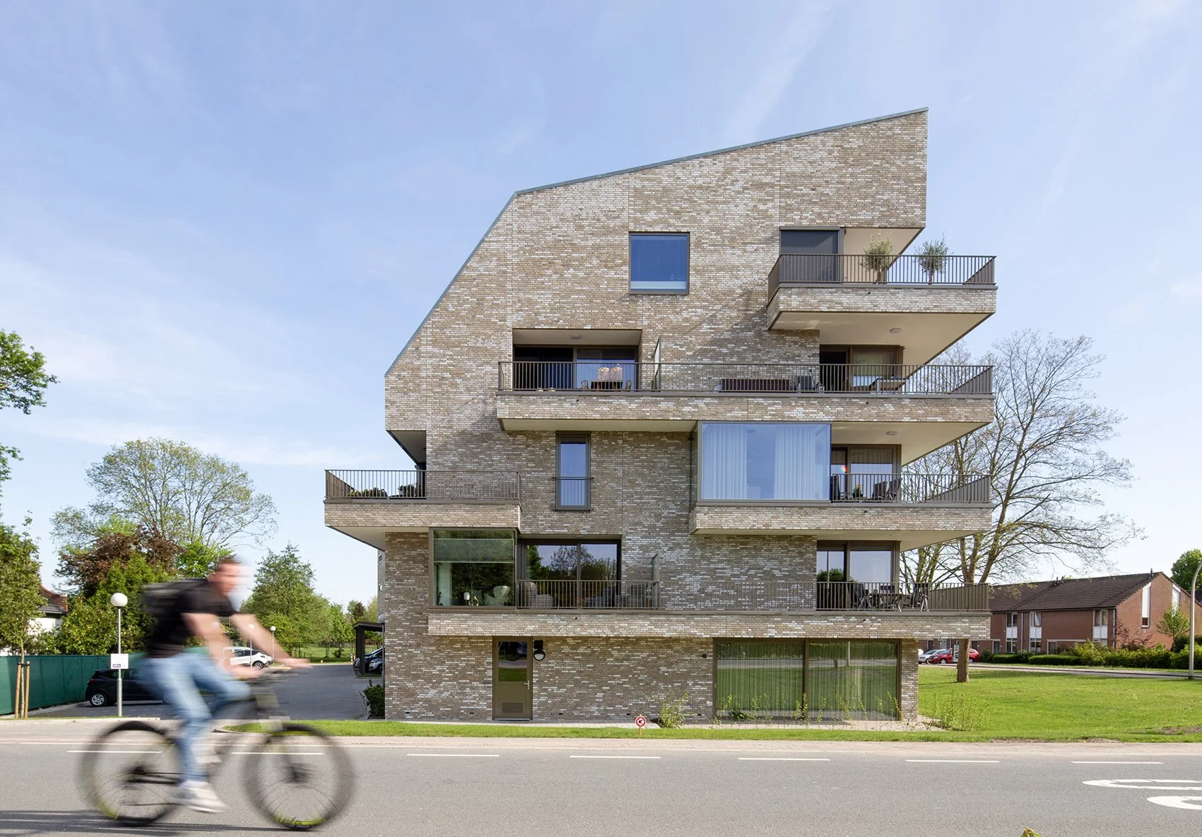 Modern vier verdiepingen tellend appartementsgebouw met balkon en glazen deuren, met bomen en blauwe hemel op de achtergrond, en een fietser die voorbij rijdt