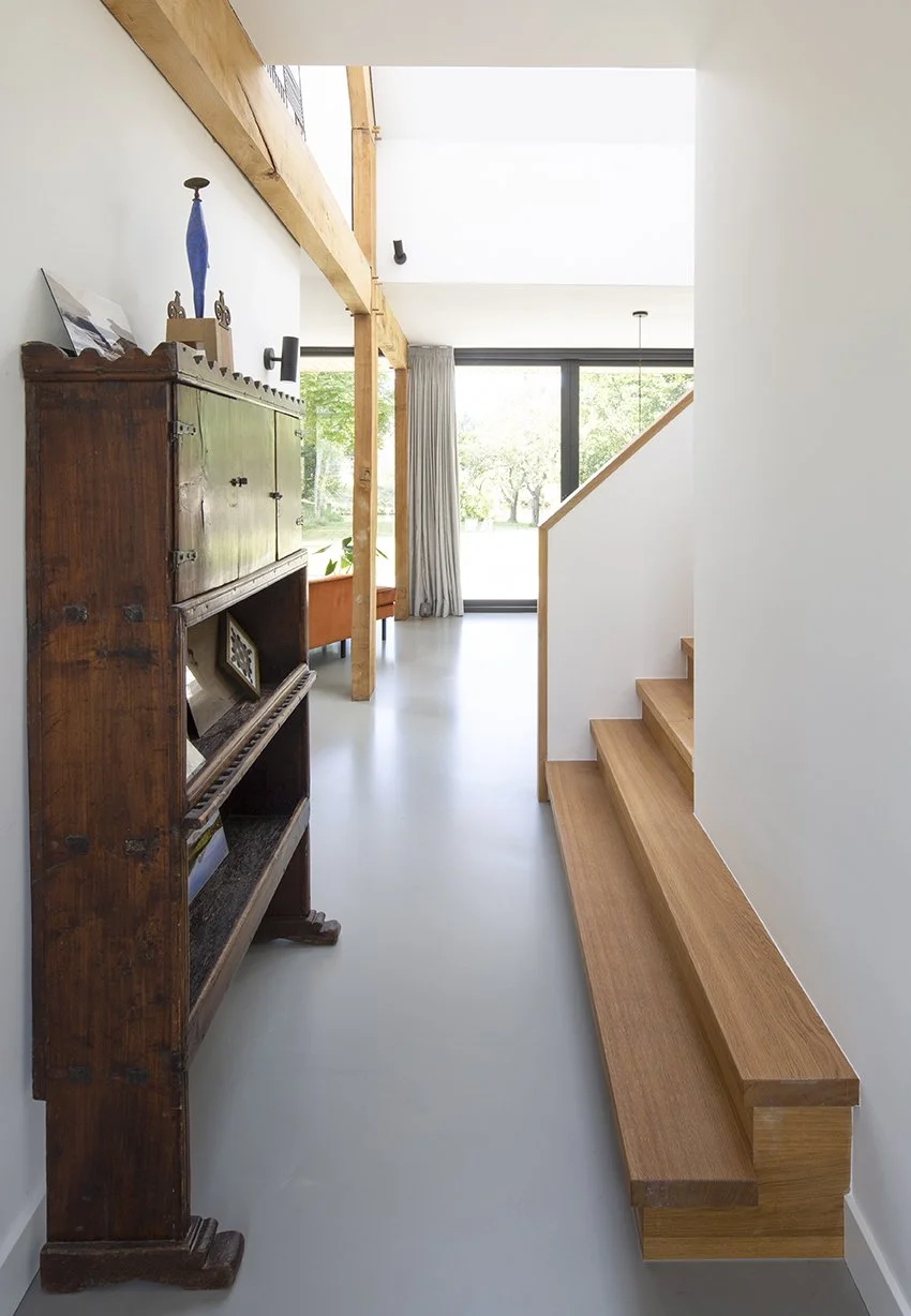Interieur van een modern huis met een houten trap, een oude houten kast en grote ramen die uitkijken op een groene tuin.