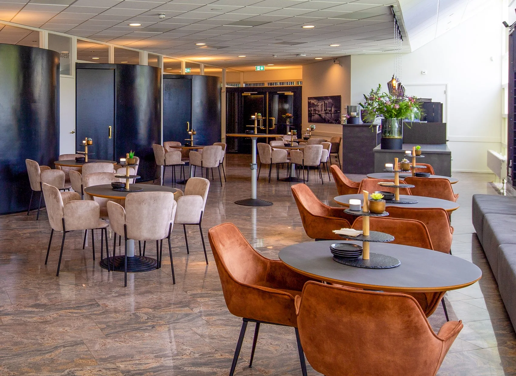 Een moderne restaurant of café met ronde tafels en comfortabele eetkamerstoelen, decoratief met bloemen en kaarsen, met donkergekleurde scheidingswanden en een lichte, gezellige sfeer.