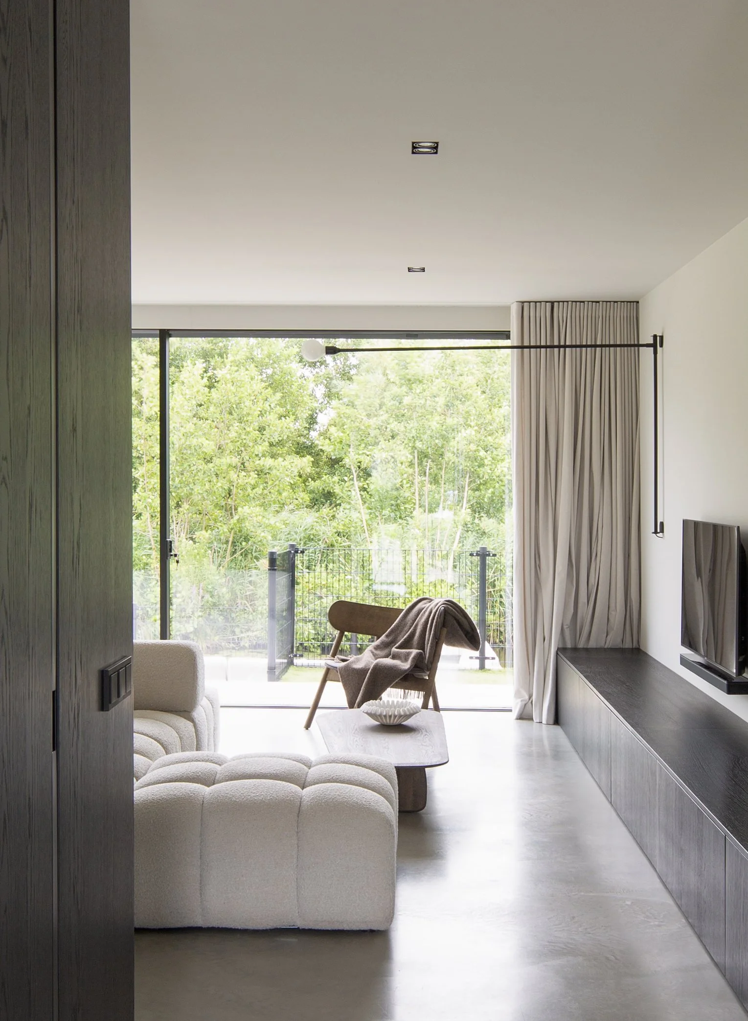 Een modern woonkamer met grote raampartijen die uitkijken op een groene tuin. Er staat een lichte bank, een comfortabele stoel met een deken, een witte salontafel met een schaal en een muurtelevisie. Gordijnen hangen langs de ramen, en het plafond he
