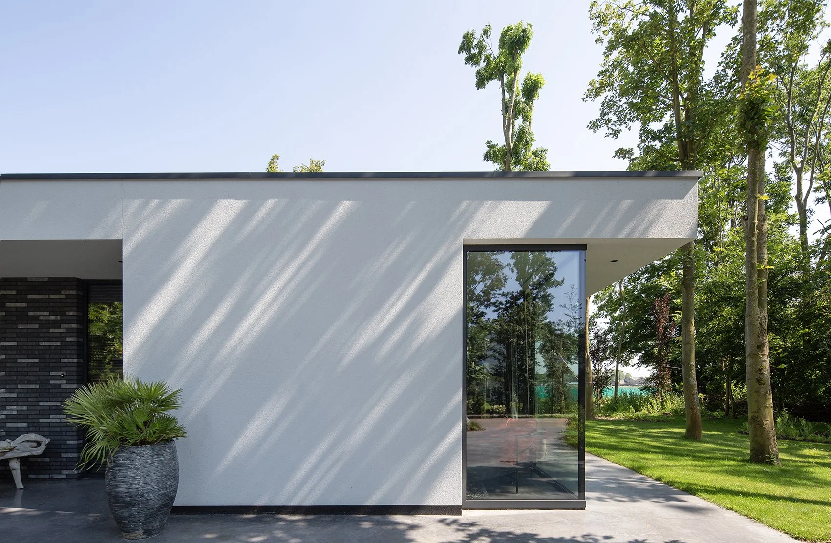 Moderne witte woning met grote glazen raampartij, omgeven door groene bomen en een gazon, met een muur van donkere bakstenen aan de linkerzijde.