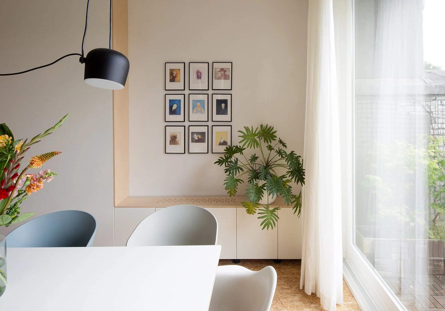 Interieur van een eetkamer met een bloemenvaas, een moderne muurdecoratie met negen fotolijsten, een grote plant, een witte tafel en stoelen, een raam met witte gordijnen en uitzicht op een patio.
