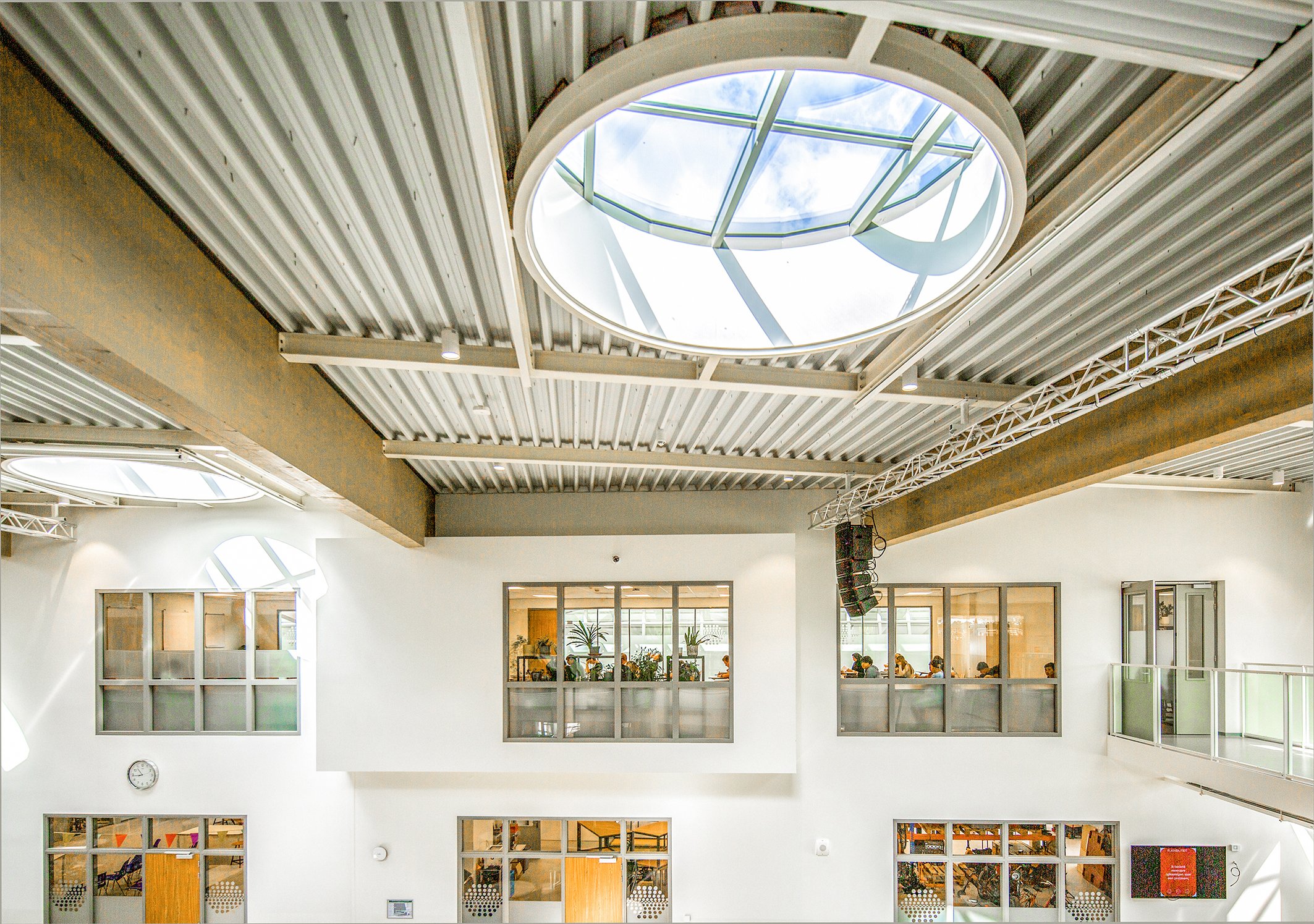 Binnenkant van een modern gebouw met grote ronde dakramen, glas en metalen constructies, met mensen in een ruimte die op de onderste verdieping zit.