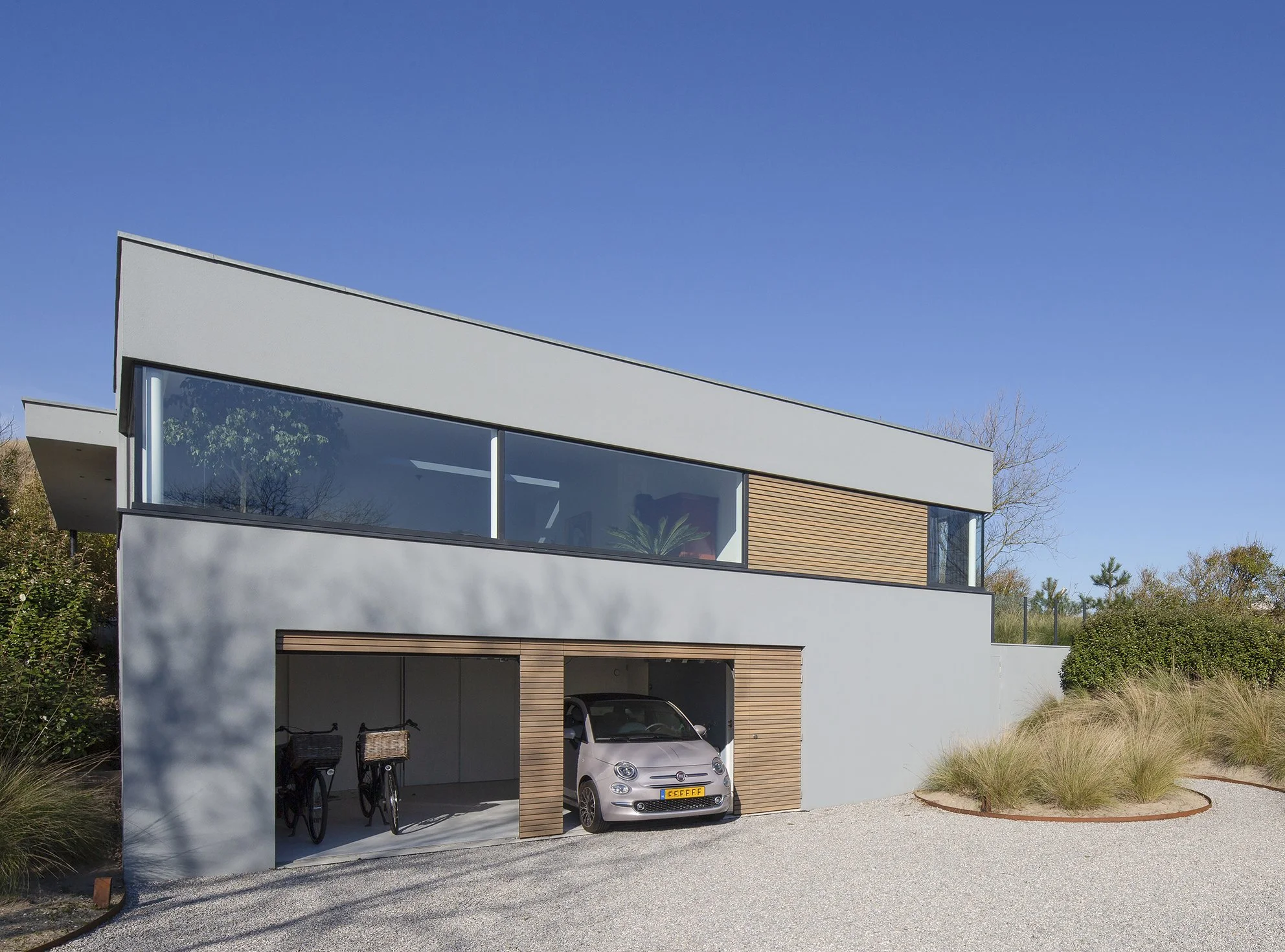 Moderne witte woning met grote ramen, houten accenten, parkeergarage met een witte Fiat en fietsen, tuin met gras en struiken onder een heldere blauwe lucht.
