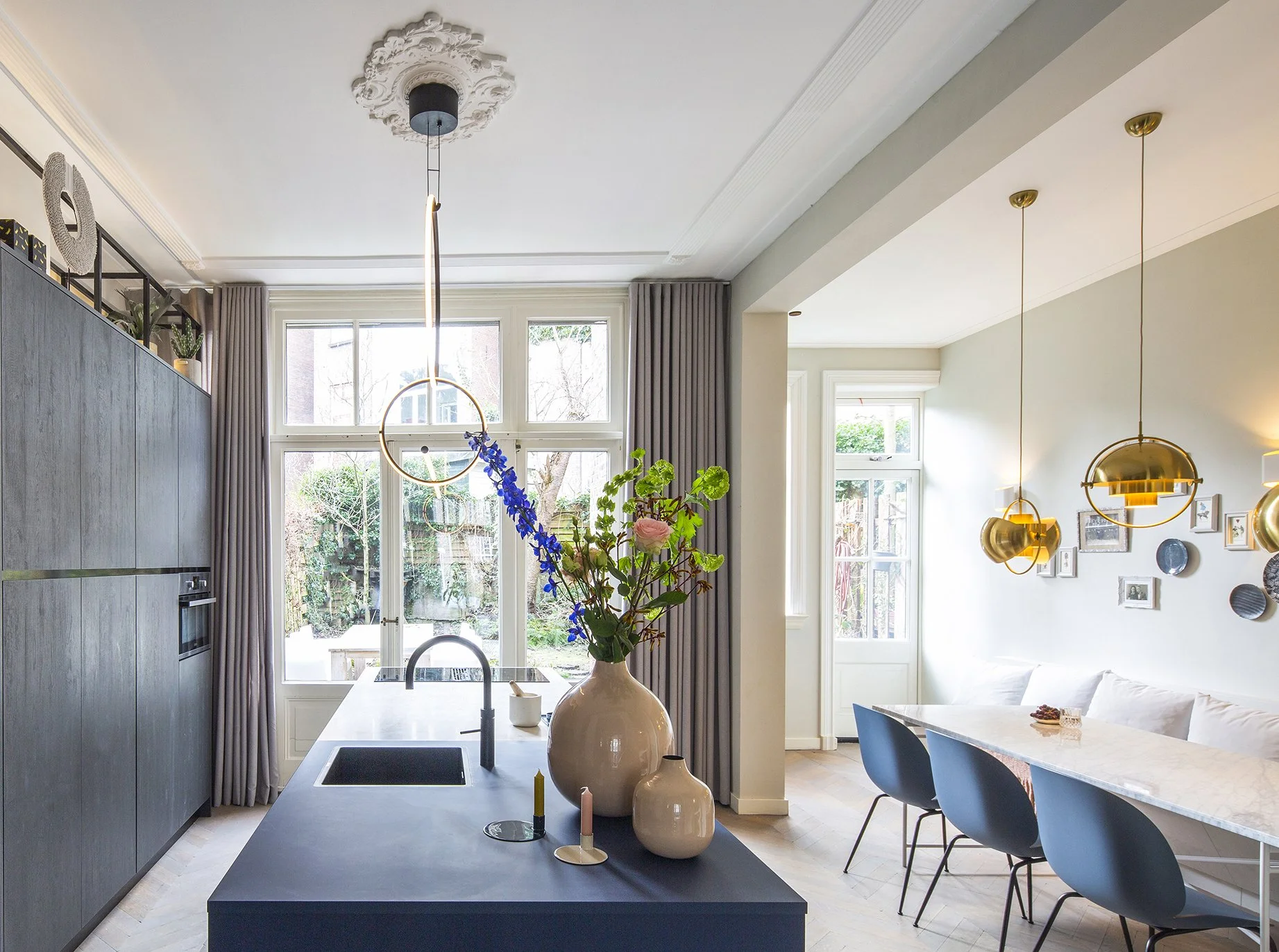 Interieur van een moderne keuken en eetkamer met grote ramen, een eiland met een gootsteen, decoratieve vazen met bloemen, en hanglampen, met een wand met foto- en kunstwerken.