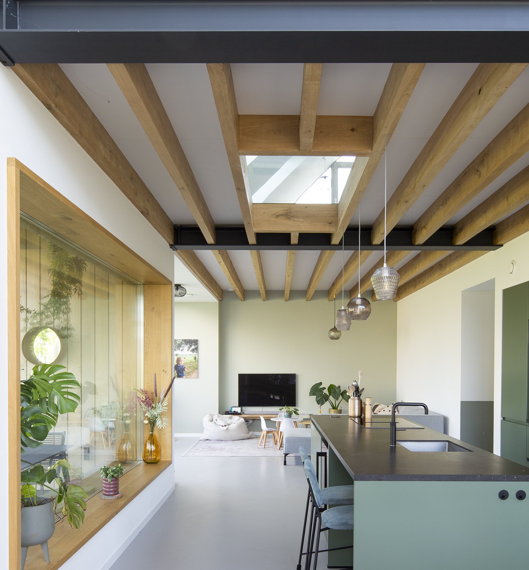 Gezicht op een moderne open keuken en woonkamer met houten plafondbalken, planten, geometrisch design, en een kleine zitruimte met een tv.