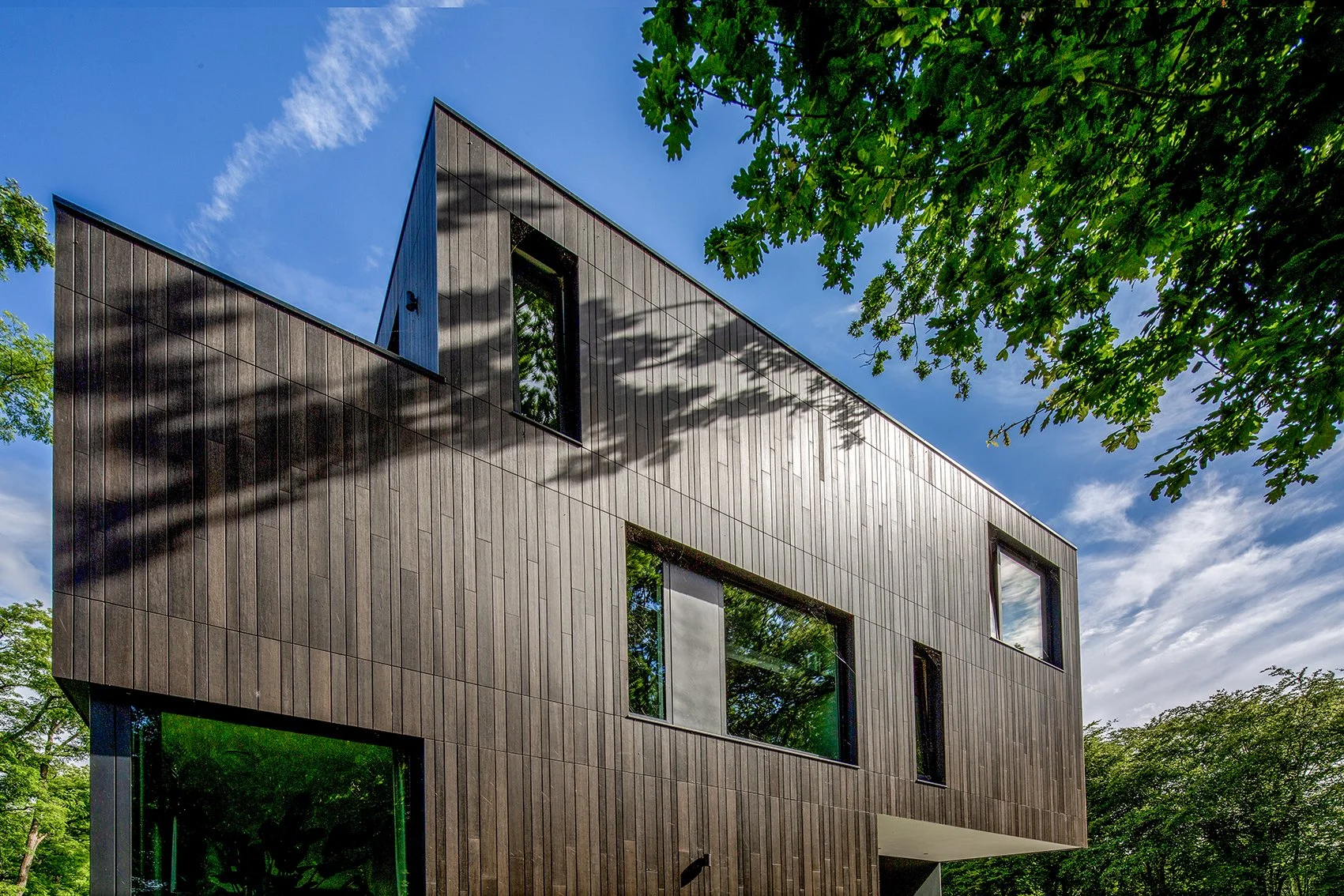 Moderne houten gebouw met grote ramen onder een blauwe hemel en groene boombladeren.