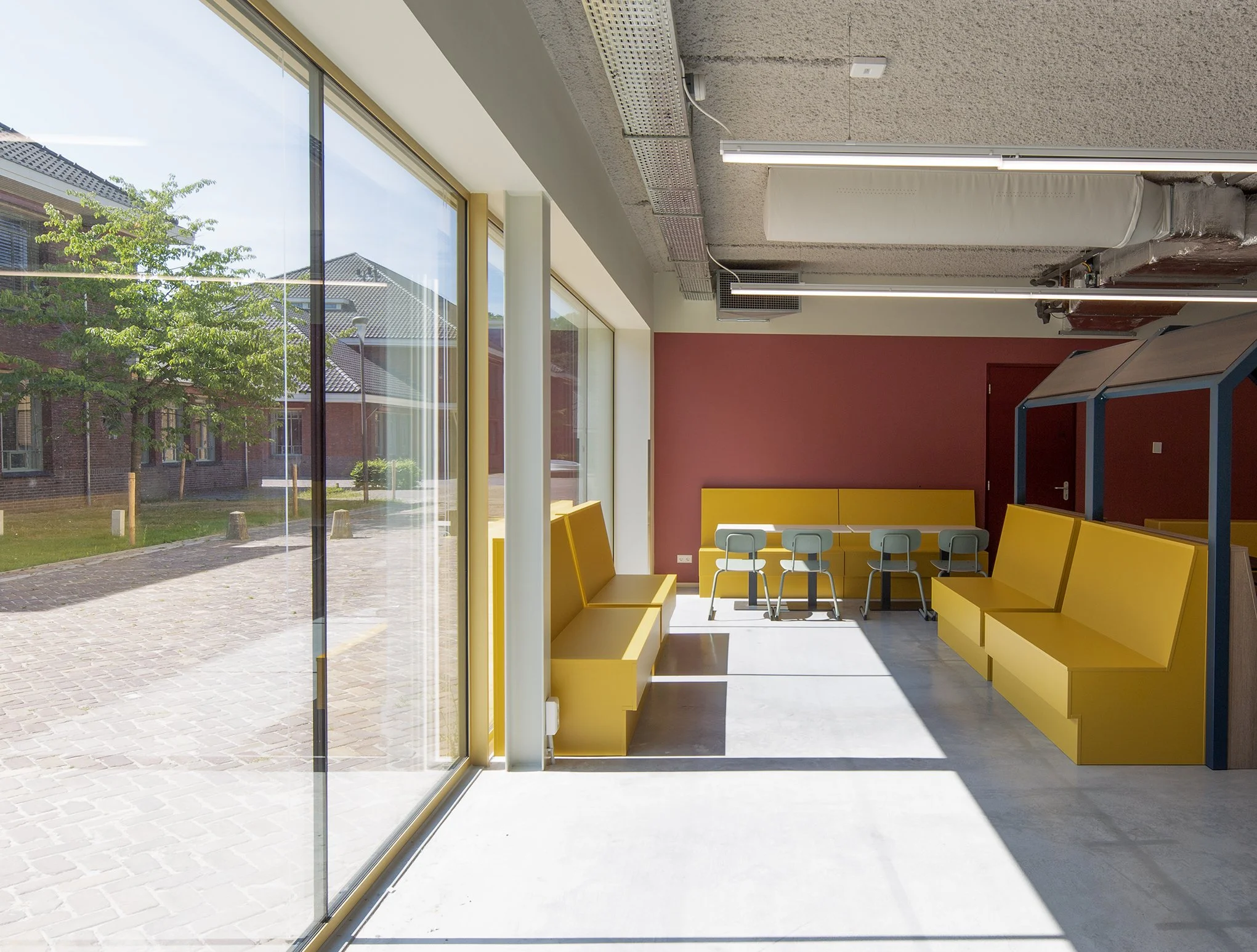 Binnenzicht van een modern interieur met gele bankjes, hoge ramen, en een muur in dieprood. Boven het hoofd hangt een fluorescentielamp en een verlaagd plafond met buizen en kabels.