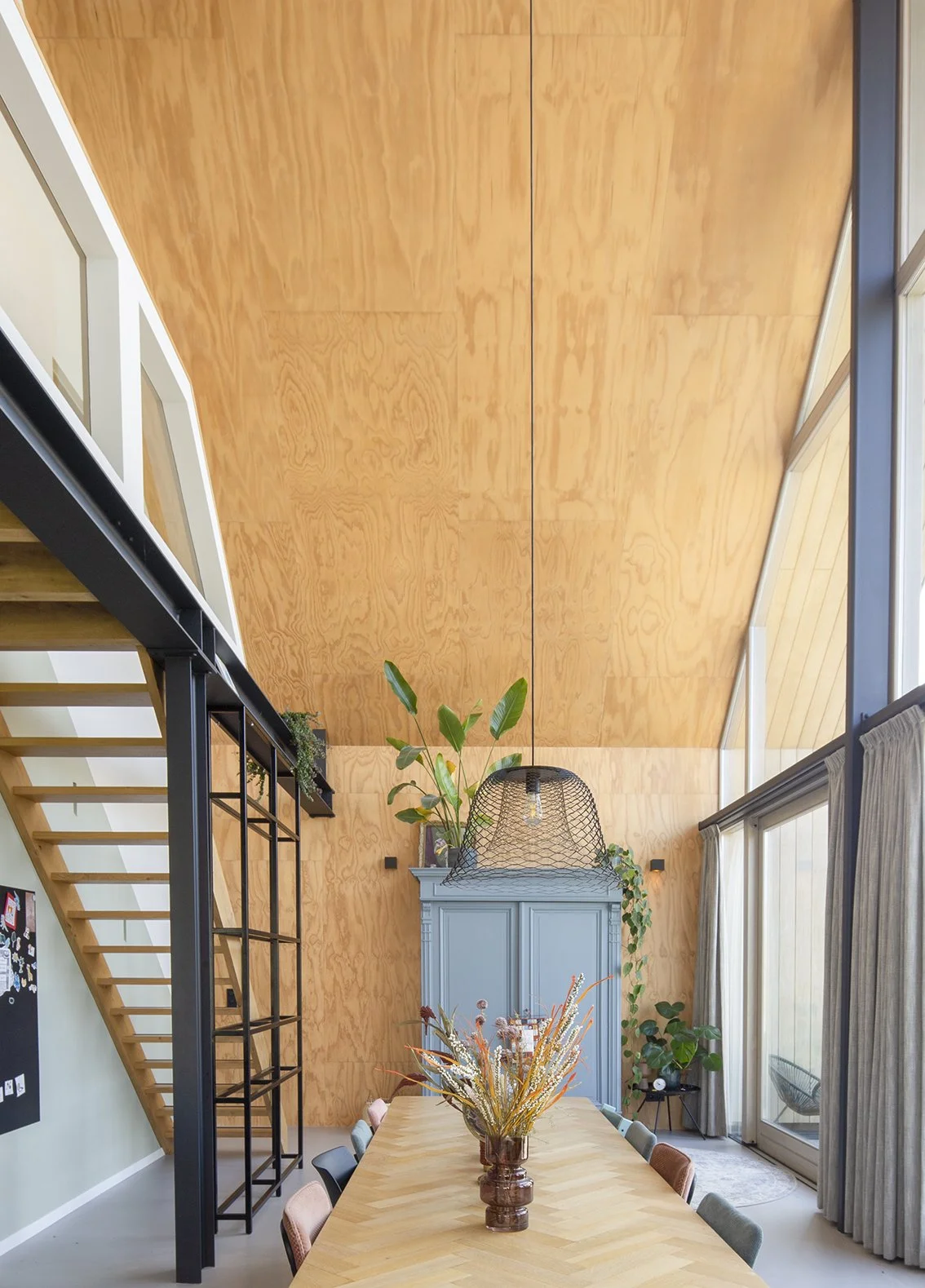 Een interieur met een hoge houten plafond, een lange tafel met een vaas bloemen, en grote ramen met gordijnen.
