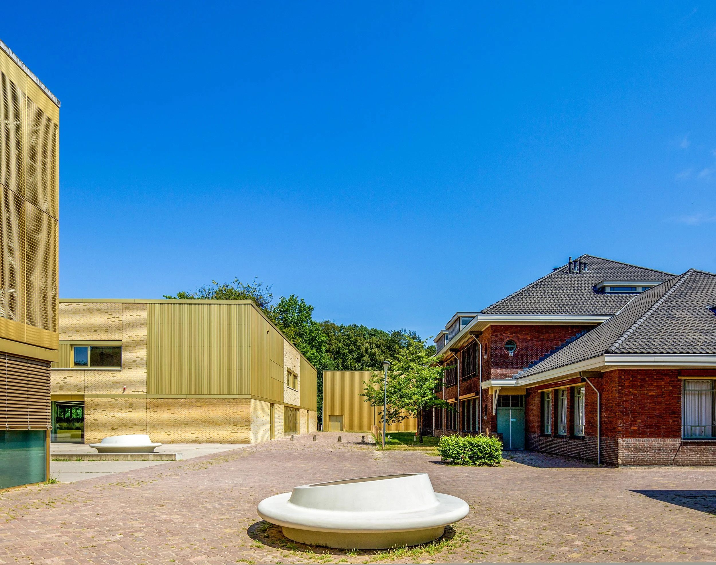 Een open binnenplaats tussen moderne gebouwen met een blauwe lucht boven. Er staan bomen, en er is een grote, witte, ronde zitplaats in het midden.