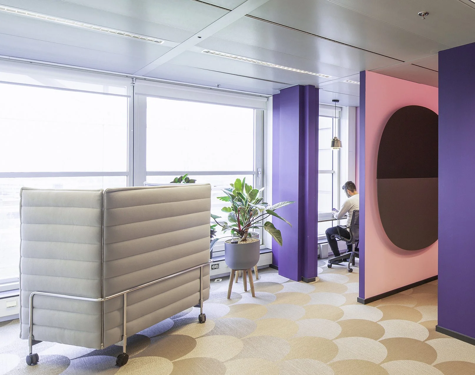Interieur van een modern kantoor met grote ramen, planten, een betonnen scheidingswand, en een man die op een bureaustoel zit.