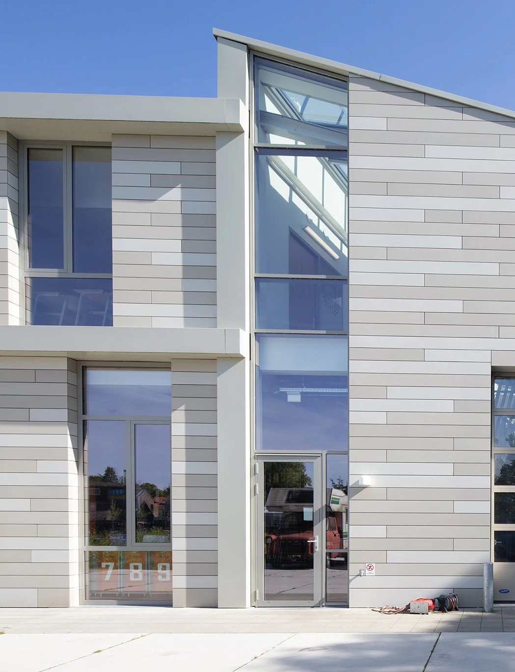 Moderne witte gebouw met grote ramen en een glazen doorgang, onder een blauw hemel.