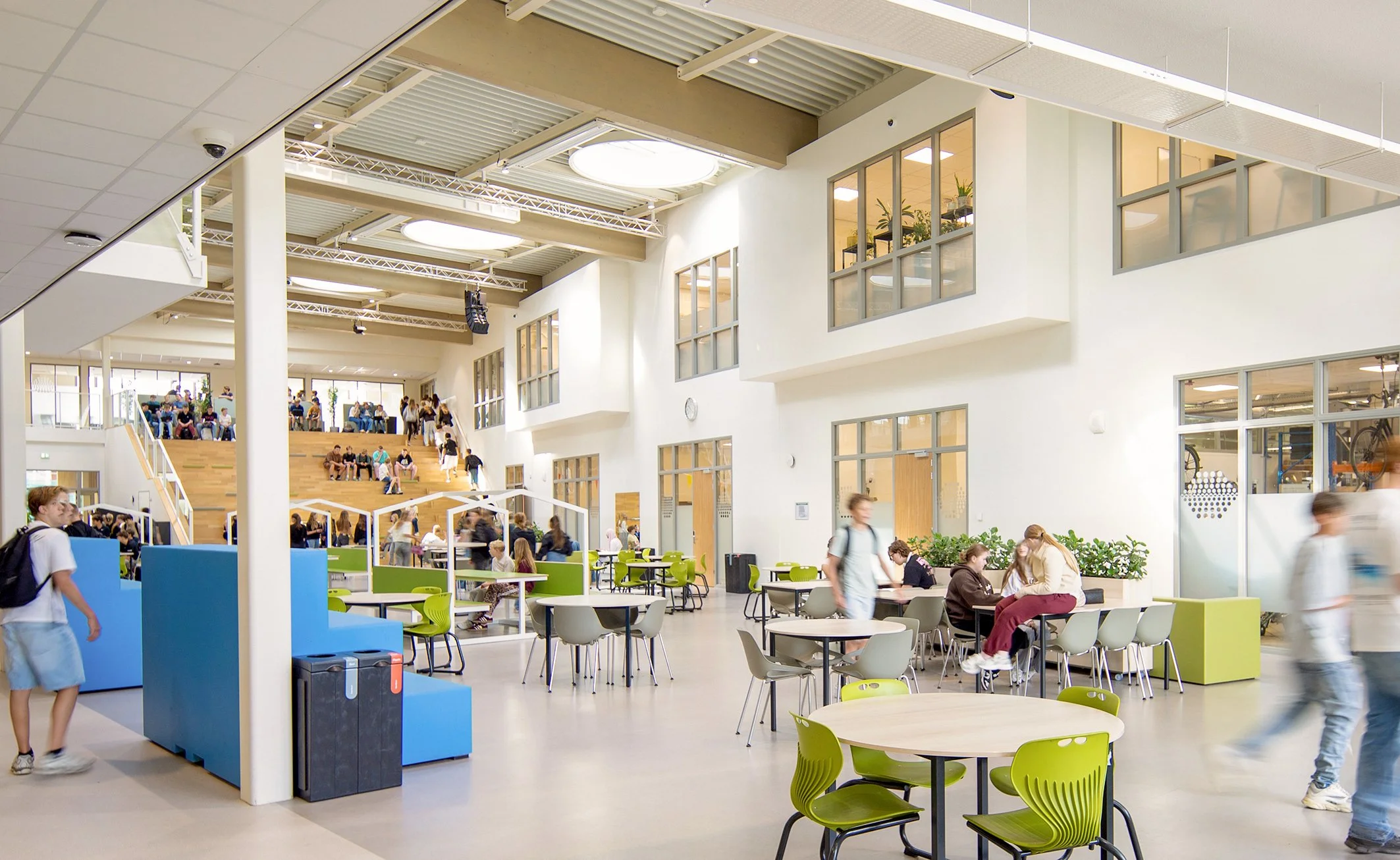 Binnenruimte van een modern station of universiteit met zitplaatsen, mensen die lopen en een verhoogd zitgedeelte met groepen mensen. Grote ramen, groene stoelen en planten.