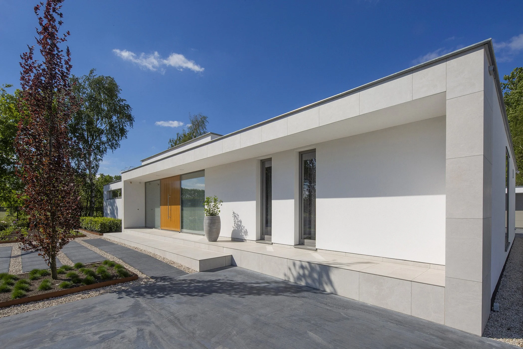 Moderne witte woning met grote ramen en een houten deur, omgeven door een tuin met bomen en planten, onder een heldere blauwe hemel.