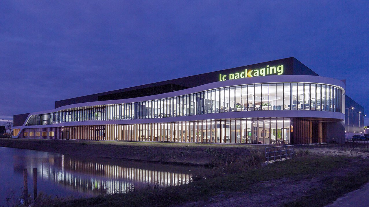 Moderne kantoorgebouw met grote glazen wanden, verlicht van binnen, boven een waterkant, met een signaal 'Le packaging' op het dak, bij avond.