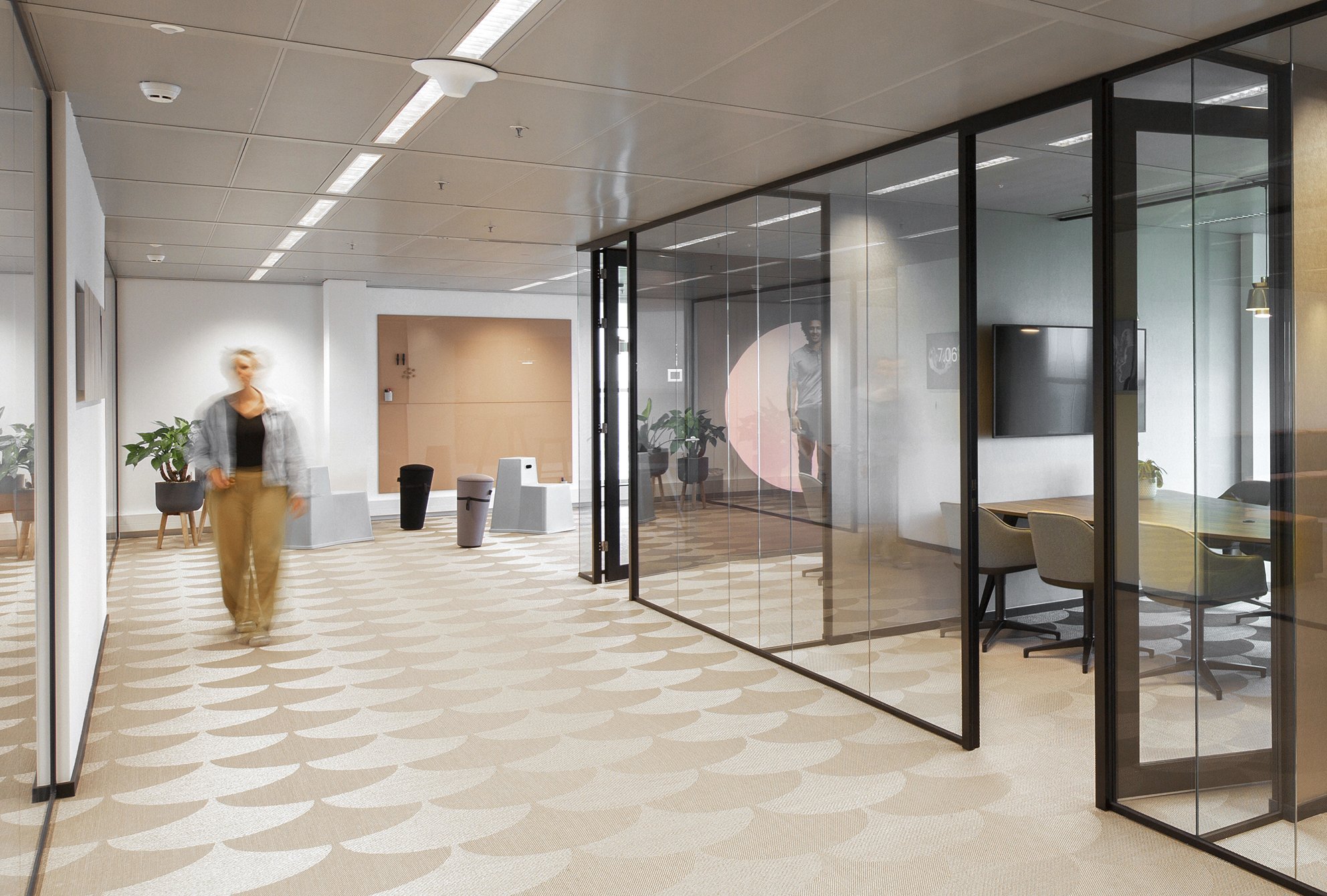 Binnenkant van een modern kantoor of lobby met een persoon wandelend op een grijze en beige vloer met een patroon. Rechterkant heeft een glazen scheidingswand met een zitgedeelte en een tv-scherm. Linkerkant is een witte muur met enkele planten.