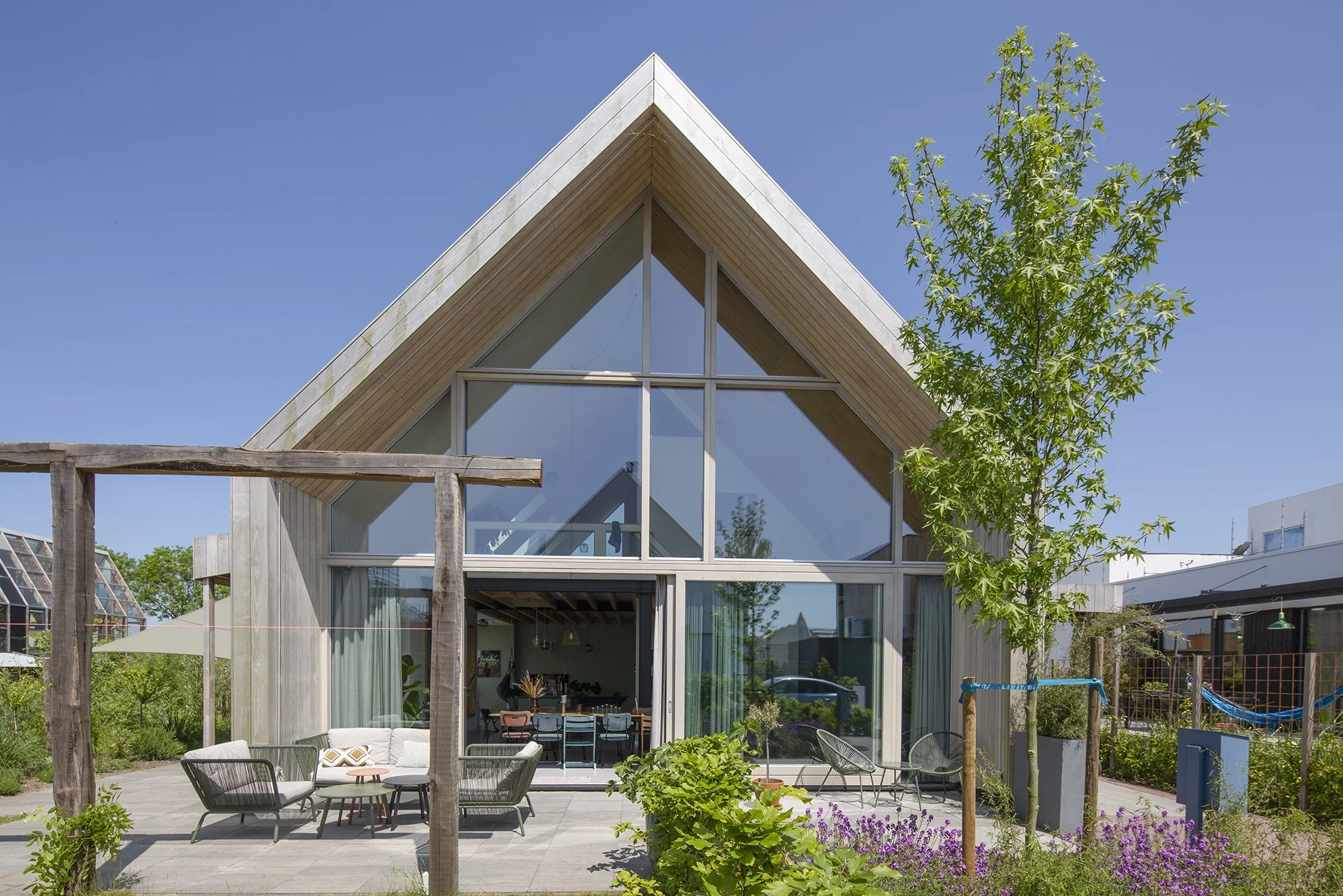 Moderne houten huis met grote glazen ramen en een tuin met zitgedeelte, terras, bomen en bloemen onder een heldere blauwe hemel.