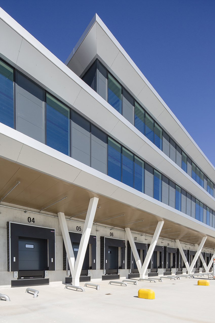 Moderne kantoorgebouw met grote ramen en een onderbouw met meerdere dockdeuren voor vrachtwagens, gebouwd op een heldere dag met blauwe lucht.
