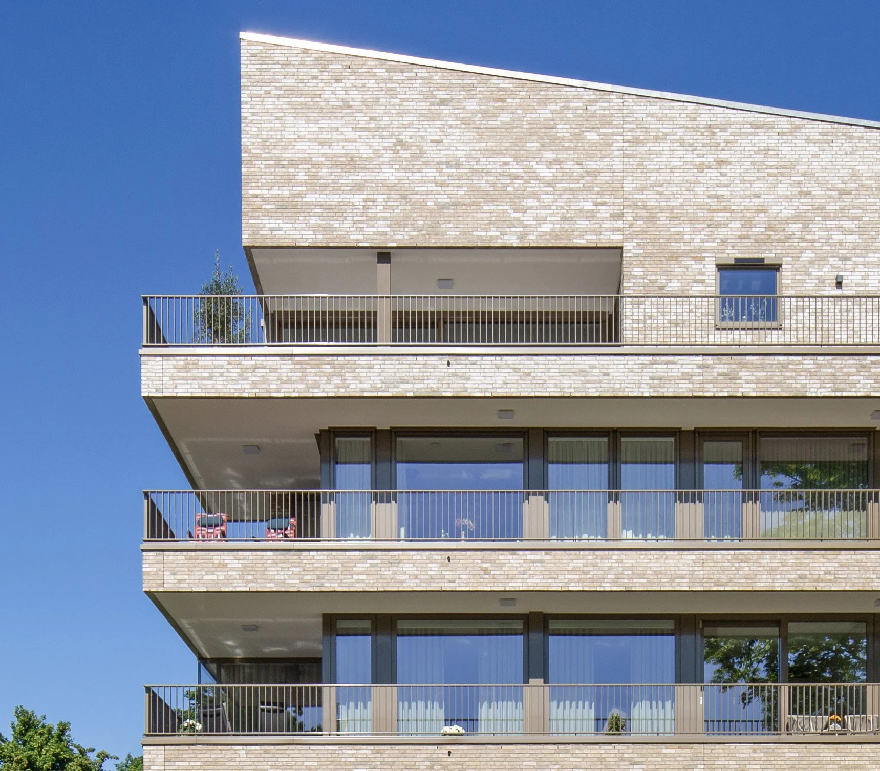 Bekijk van een modern appartementencomplex met vier verdiepingen en verschillende balkons. De gevel bestaat uit beige bakstenen en grote glazen ramen. De dag is zonnig met een heldere blauwe hemel.