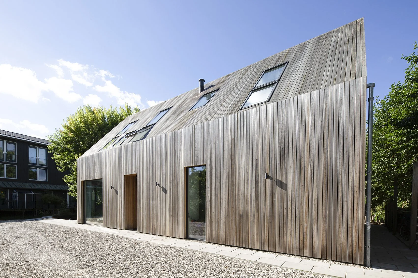 Moderne houten woning met schuine daken en grote ramen, omgeven door groene bomen onder een blauwe hemel.