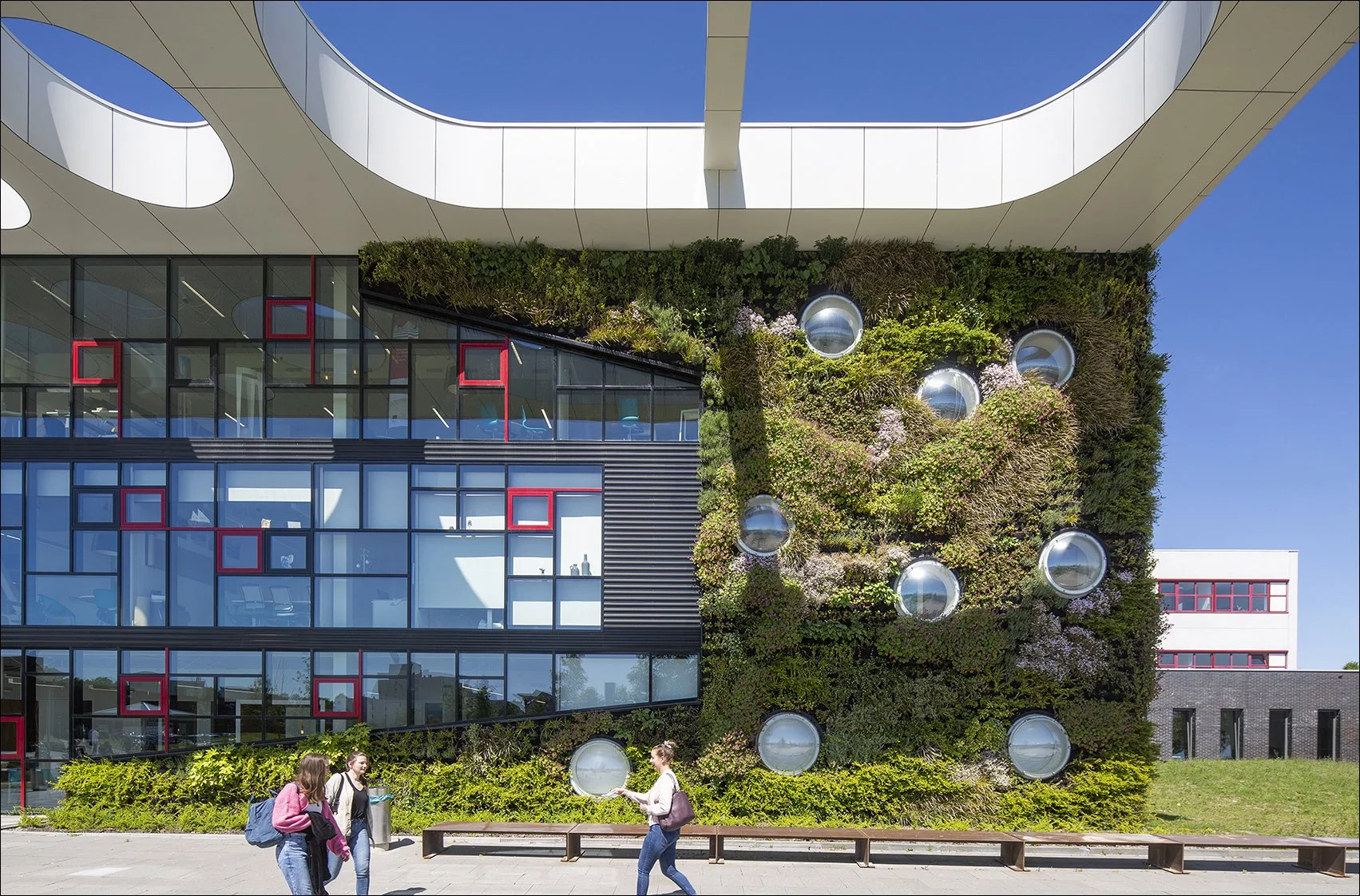 Een moderne gebouw met een groene gevel, voorzien van ronde ramen en grote ramen met rode omlijstingen, onder een witte, golvende overkapping. In de voorgrond lopen drie vrouwen die praten.