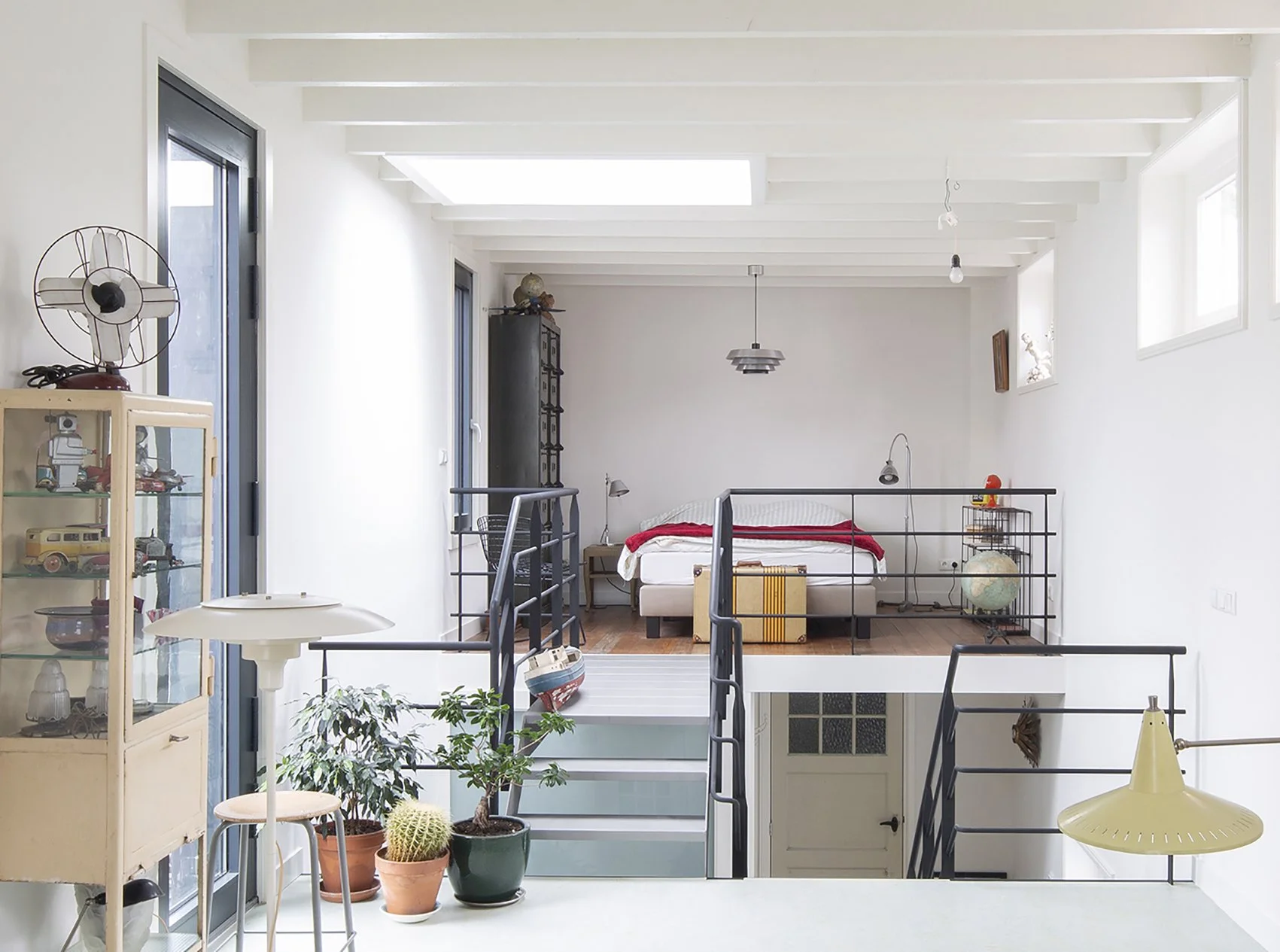 Interieur van een lichte woonruimte met een slaapkamer op de bovenverdieping, inclusief een bed met rood dekbed, een globe, planten, vintage decoratie en een glazen vitrinekast met speelgoed.