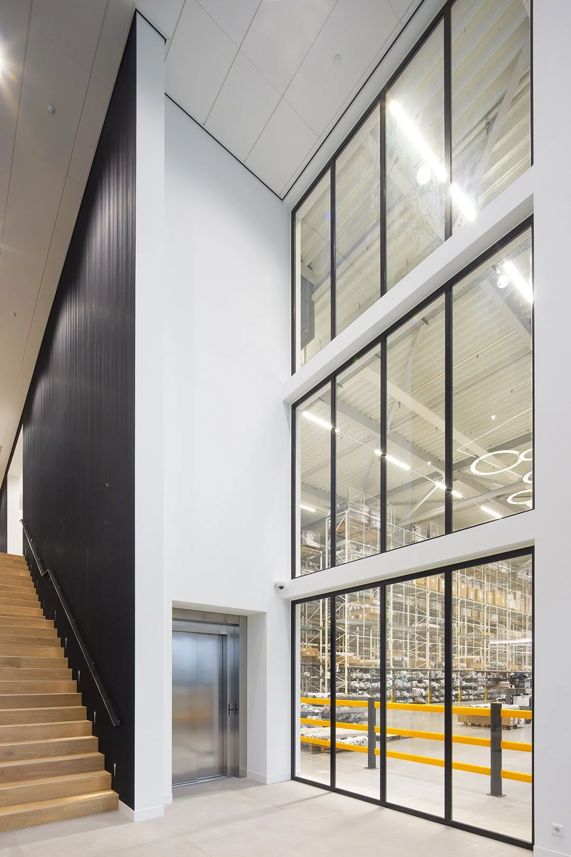 Interieur van een modern industrieel gebouw met grote ramen, trappen en een opbergruimte met stellingen en opgeslagen goederen.