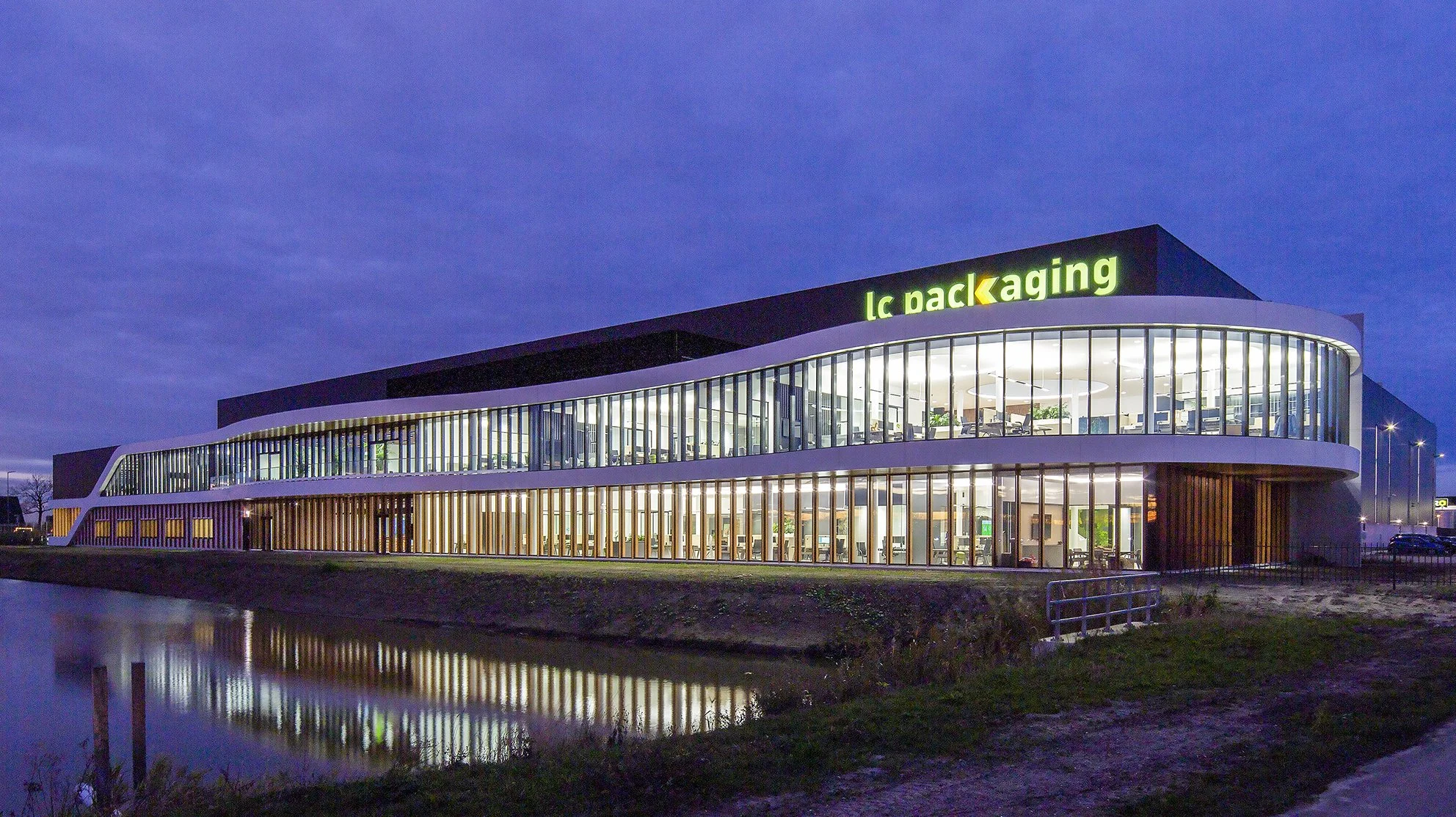 Moderne kantoorgebouw met veel glas en verlichting, met de tekst 'lc packaging' op het dak, bij schemering, naast een waterpartij.