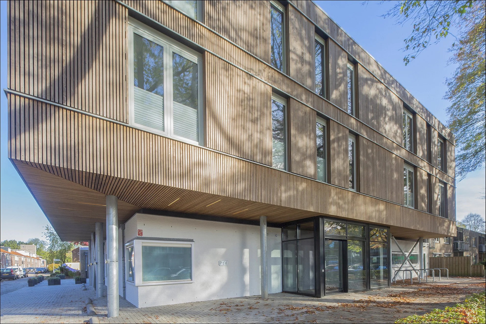 Modern gebouw met houten gevel en grote ramen, onderste verdieping met winkelruimte, omliggende straat met parkeerplaatsen en bomen.