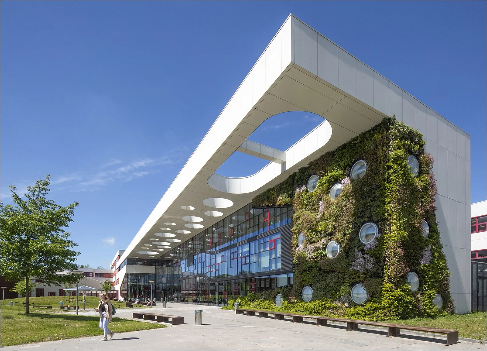 Modern gebouw met groene gevel, grote ronde ramen en een witte overkapping met cirkelvormige uitsparingen, onder een blauwe hemel.