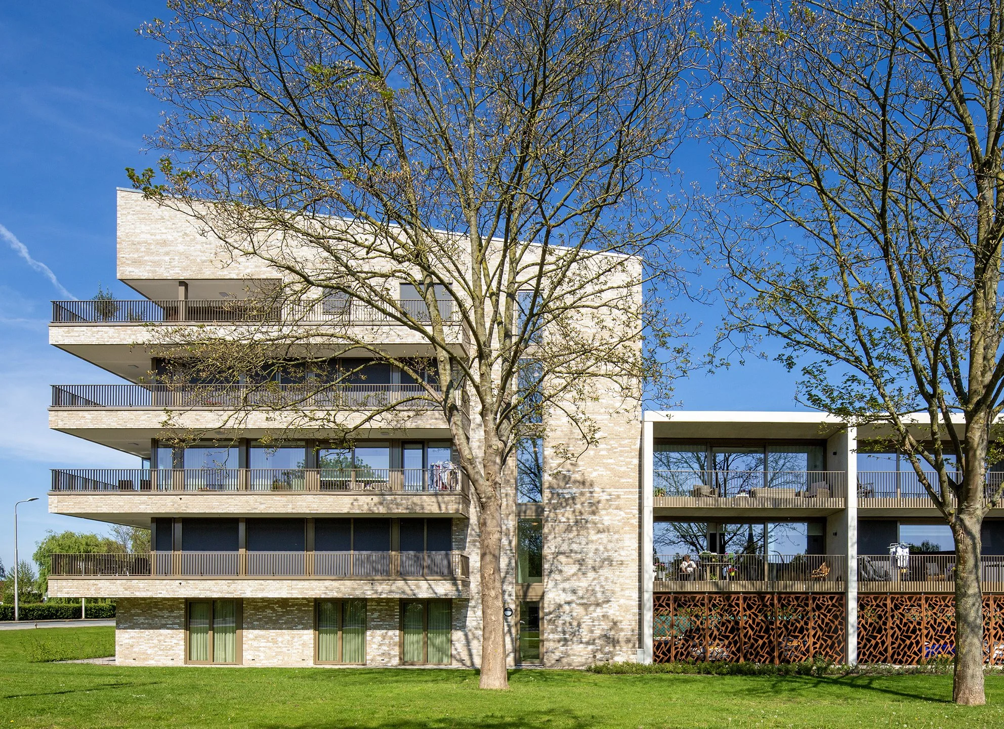 Moderne vijf verdiepingen tellende appartementencomplex met balkons en grote ramen, omringd door groene grasvelden en twee bomen, onder een heldere blauwe lucht.