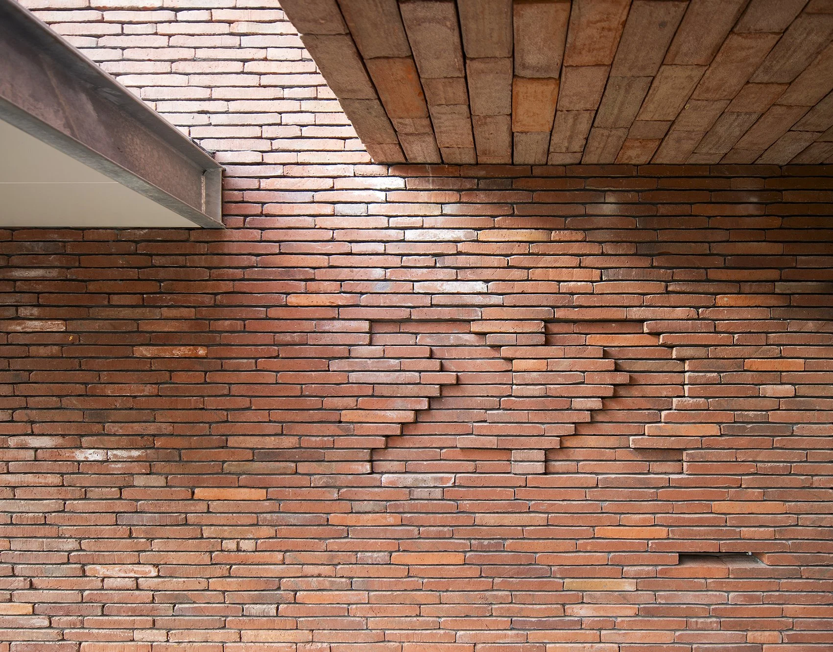 Close-up van een bakstenen muur met een afgeschuinde rand, weergegeven van onderaf met een duidelijke scheiding tussen het bakstenen deel en het plafond en een klein deel van een metalen afwerking zichtbaar.