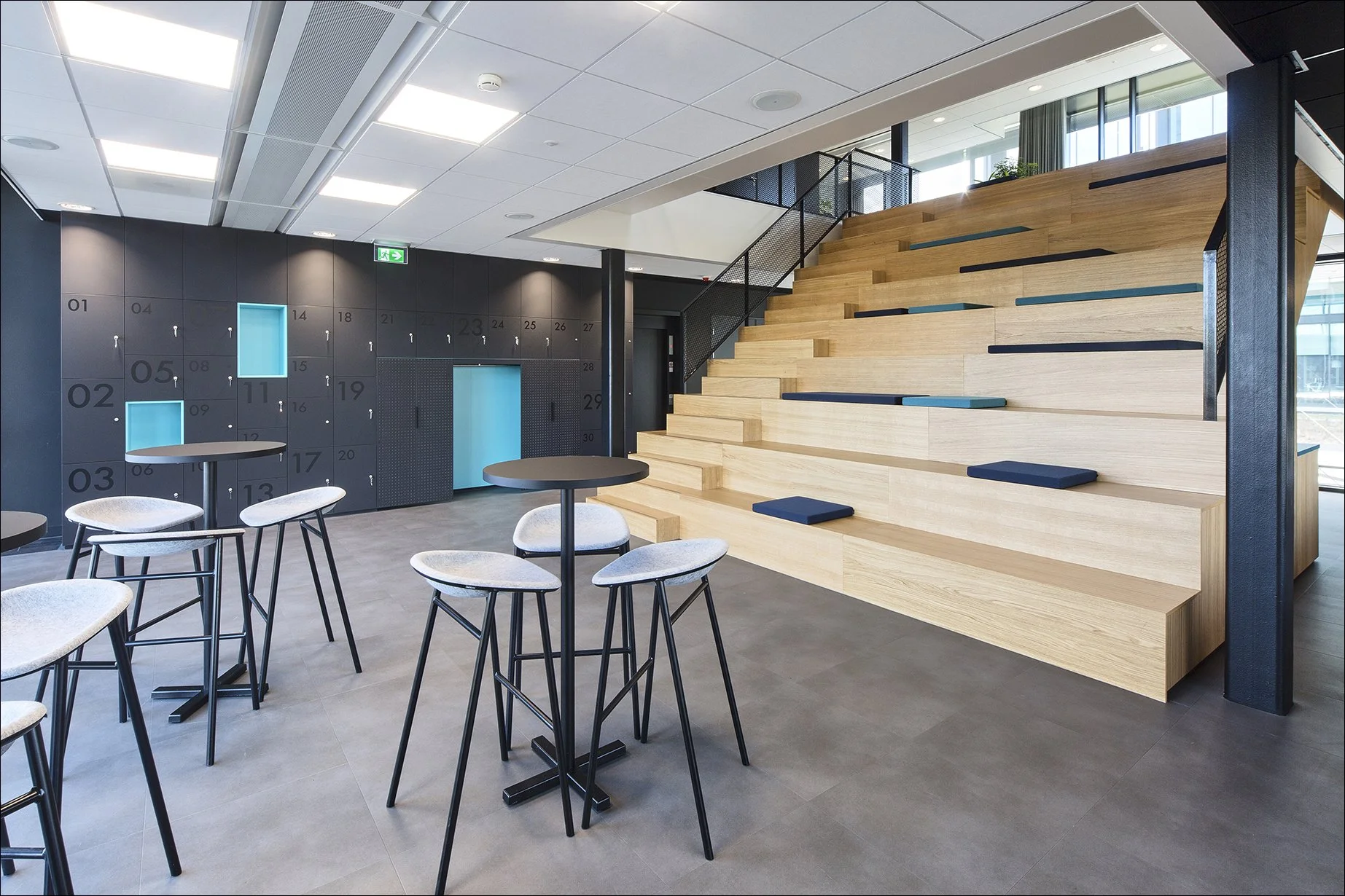 Modern interieur met hoge tafel en stoelen, zwarte lockers met blauwe accenten en een houten tribune met kussens, grote ramen en een plafond met strakke verlichting.
