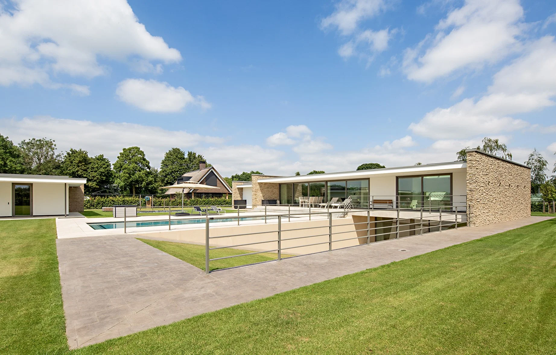 Moderne woning met groot terras en zwembad, omgeven door groene tuin, onder een blauwe hemel met enkele witte wolken gefotografeerd door architectuurfotograaf John van Groenedaal.