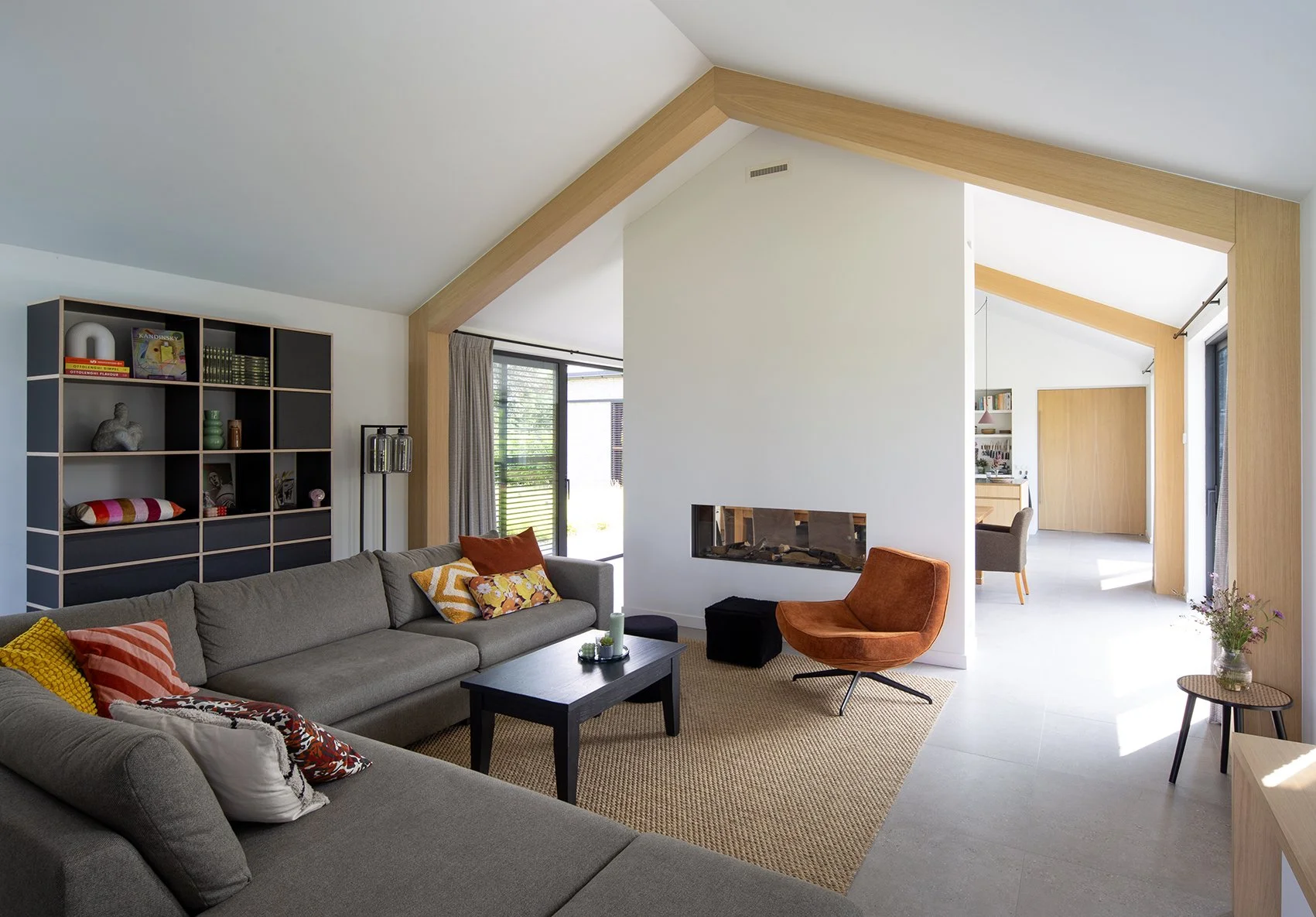 Een modern woonhuis woonkamer met een grote grijze bank met gekleurde kussens, een houten salontafel, een oranje fauteuil, een wandrek met decoratieve items en boeken, grote ramen met gordijnen, een gashaard, en een open verbinding naar een eetkamer 