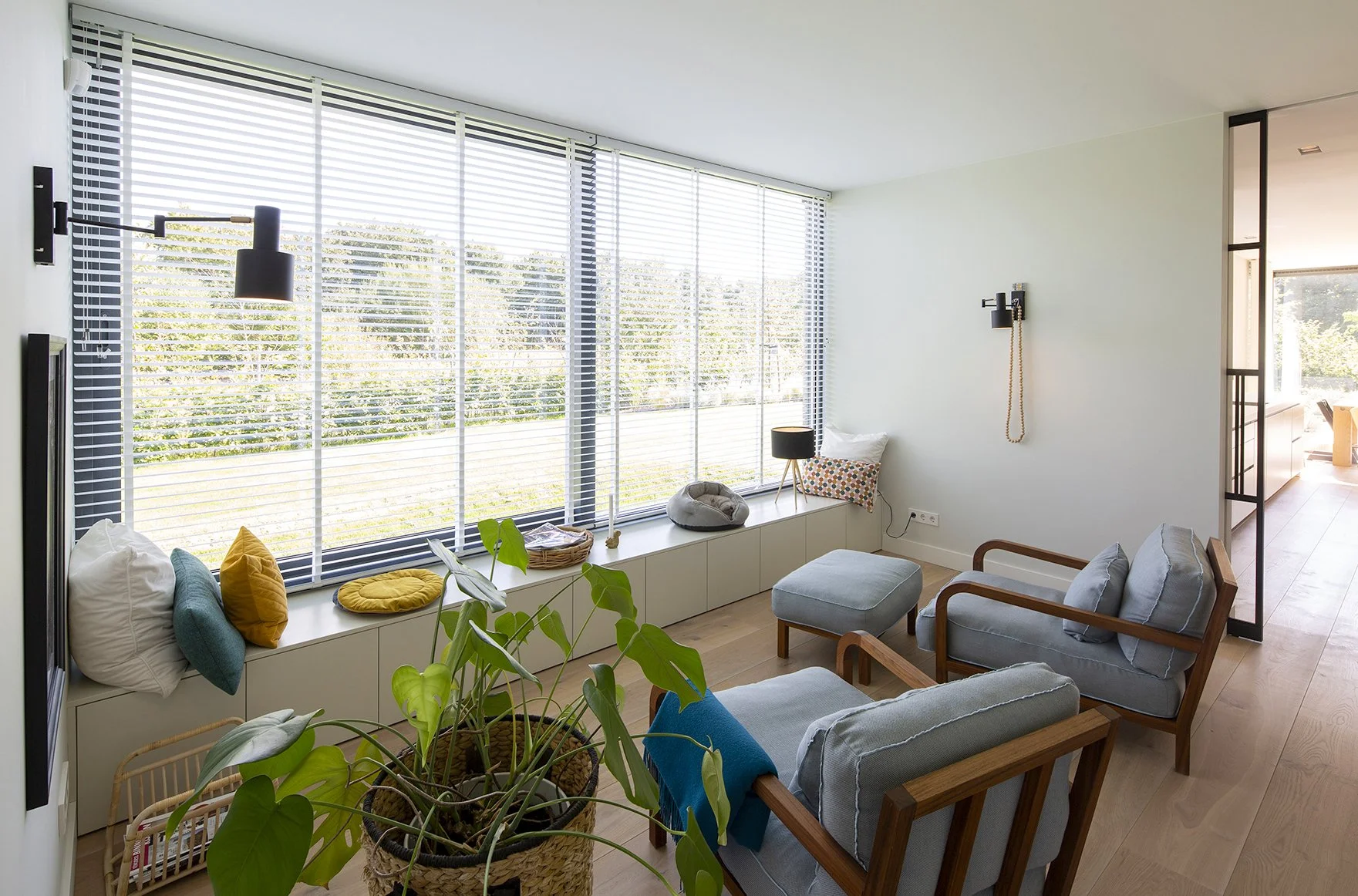 Woonkamer met grote ramen, planten, bank en fauteuils, decoratieve kussens, lezen lamp en houten vloer.