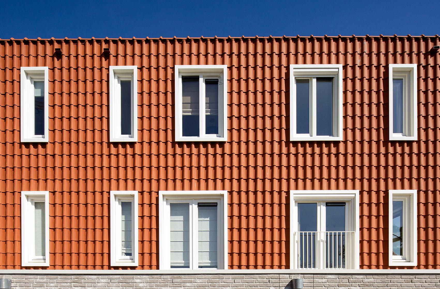 Voorgevel van een gebouw met rode dakpannen en wit omlijste ramen, inclusief een balkon en een blauwe hemel.