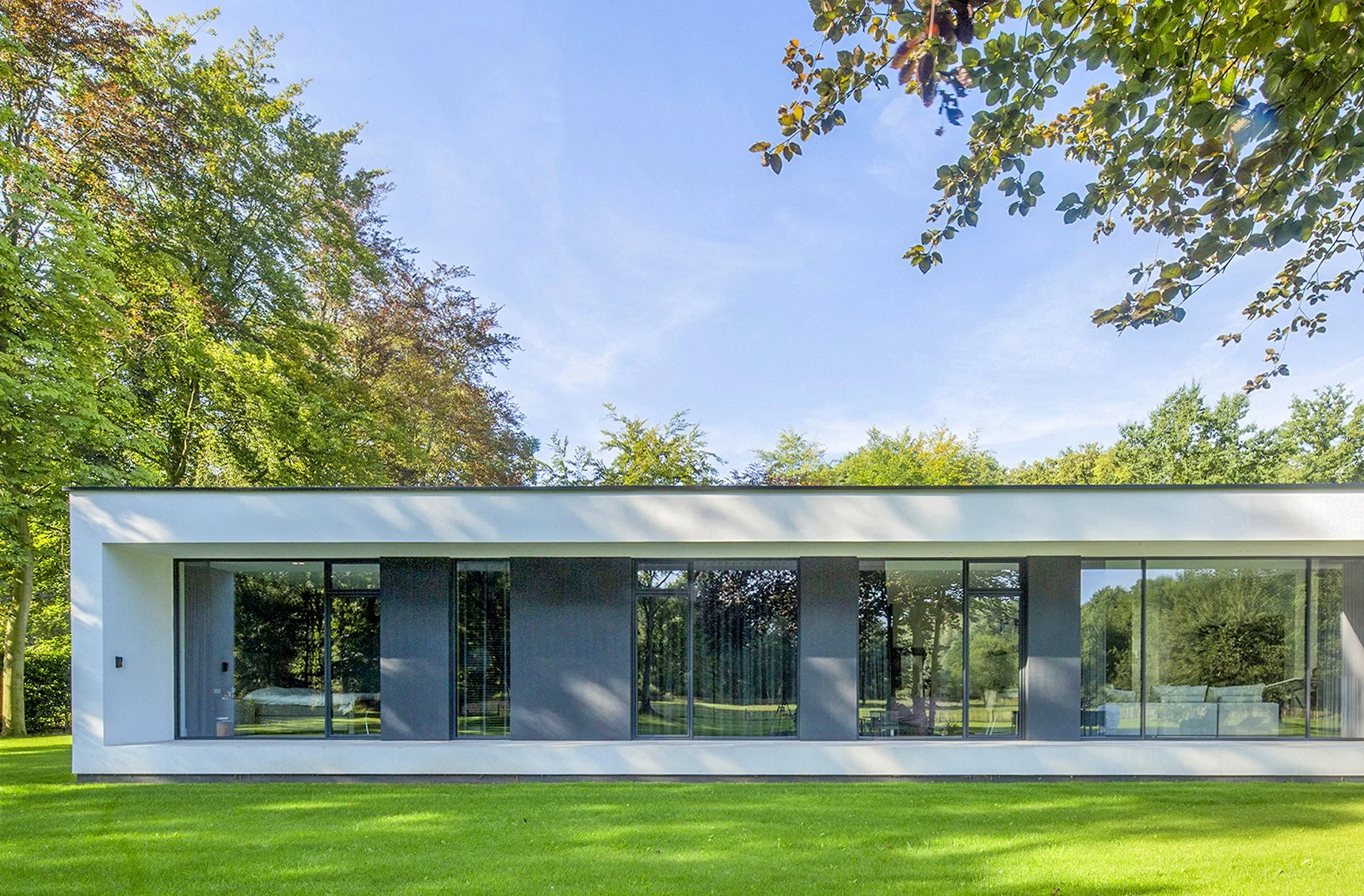 Modern witte woning met grote glazen ramen, omgeven door groen gras en bomen, onder een blauwe hemel.