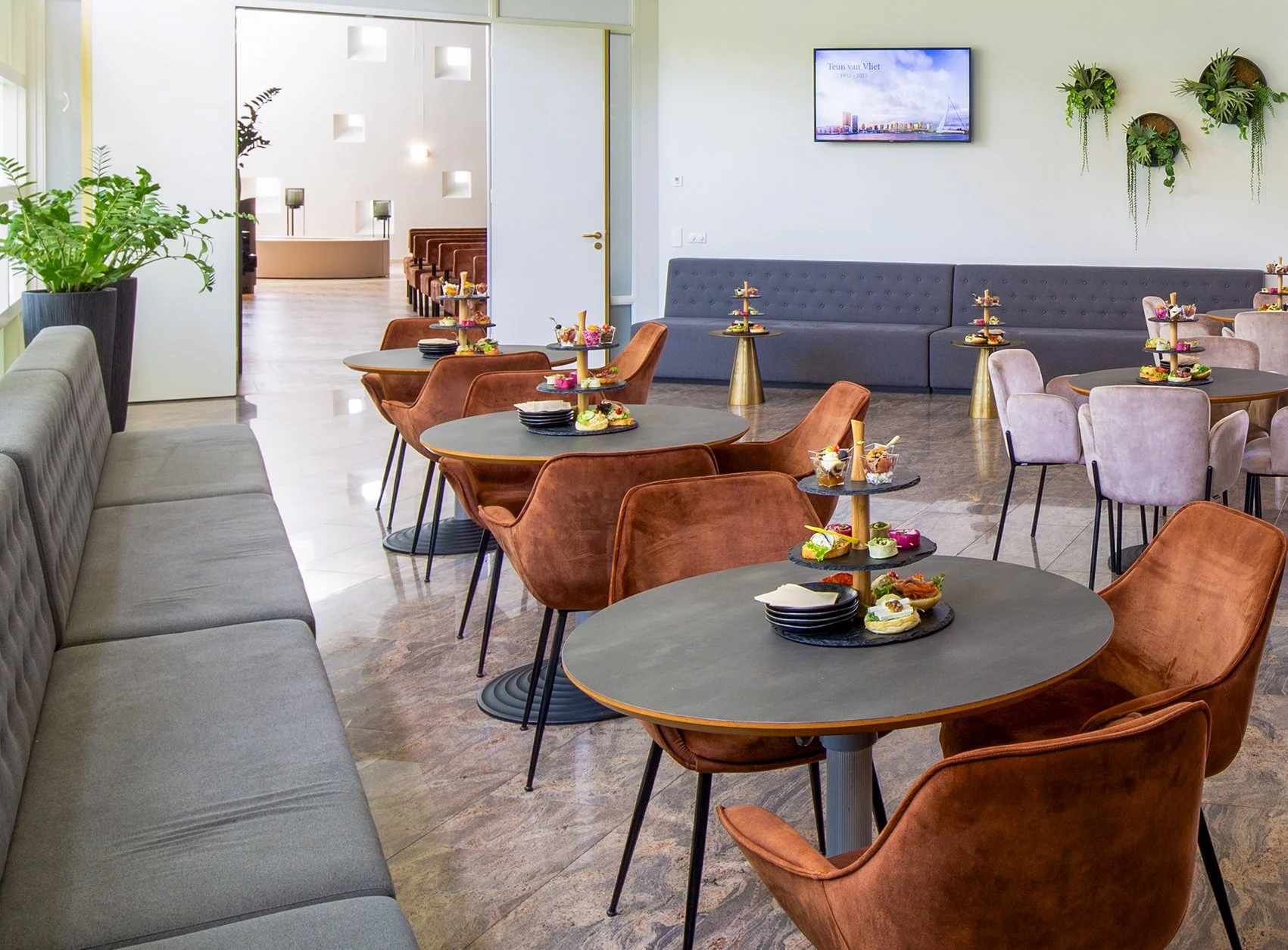 Interieur van een modern café met ronde tafels, roze en bruin beklede stoelen, een lange grijze bank en decoratieve planten aan de muur.