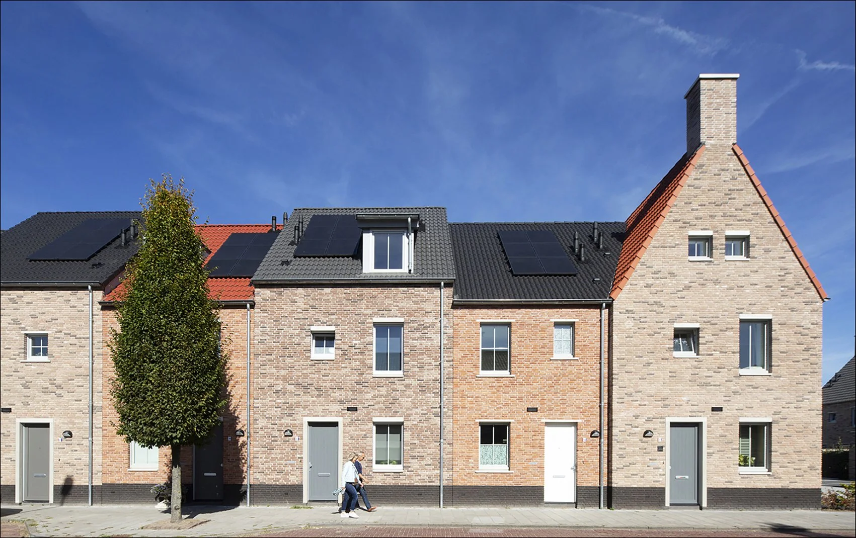 Rijen rijtjeshuizen met diverse kleuren bakstenen en zonnepanelen op de daken, twee mensen wandelen langs de trottoir, onder een blauwe lucht.