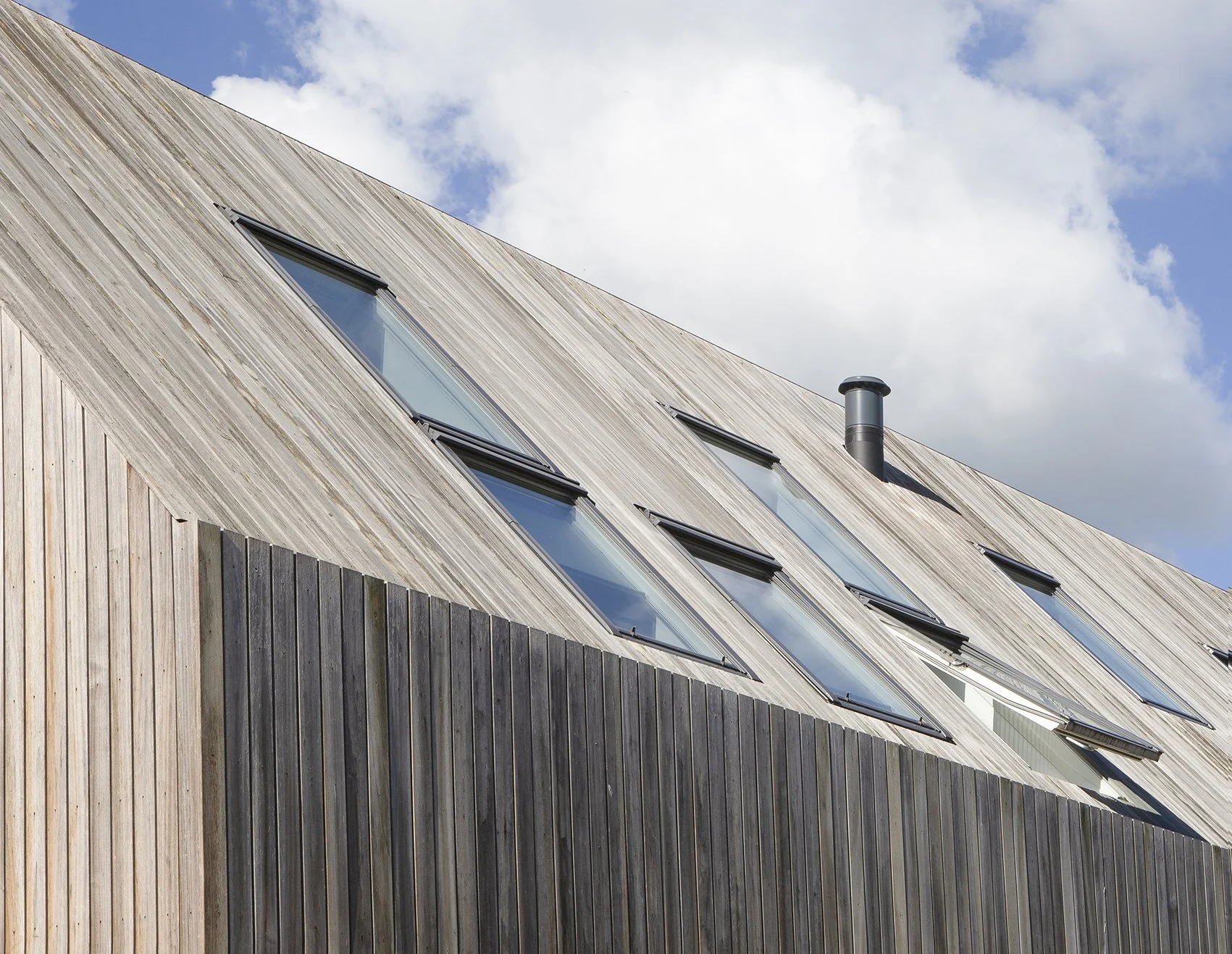Een modern gebouw met een斌dschuin hellend houten dak en grote raamluiken, onder een blauwe lucht met witte wolken.