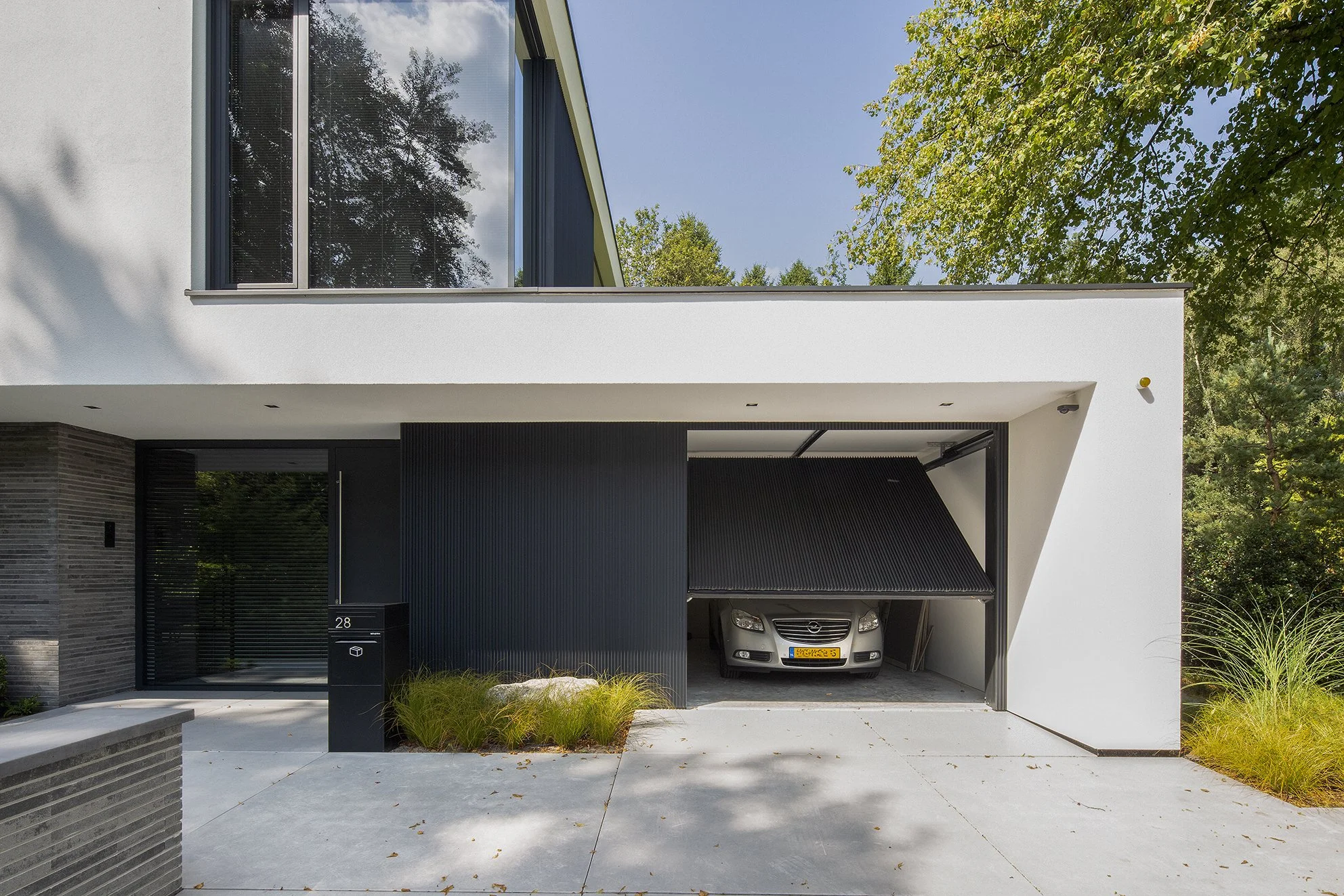 Moderne witte woning met grote ramen, een afdak voor de garage met een auto erin, groene tuin en bomen eromheen.