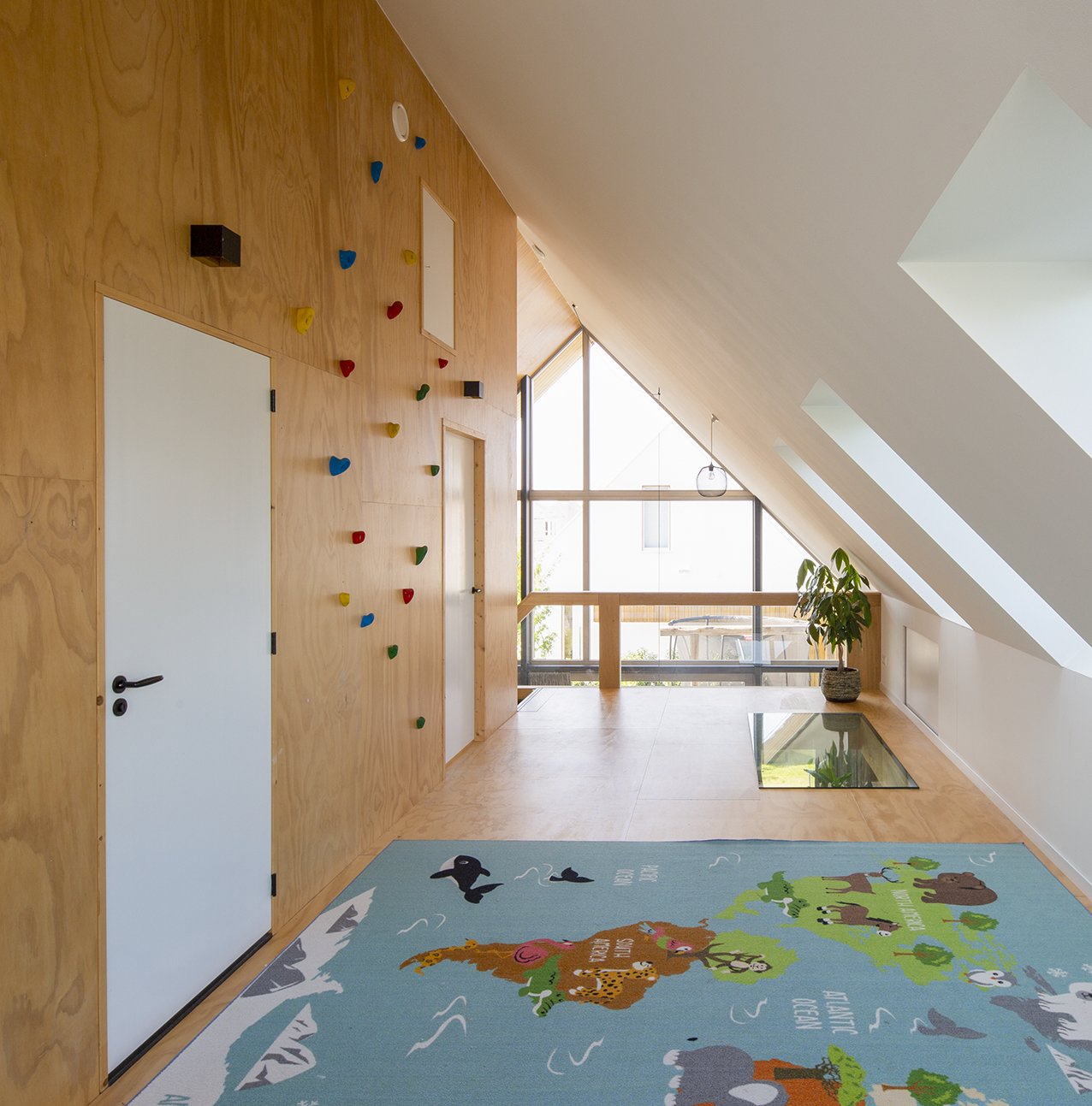 Interieur van een kamer met houten accentmuur, klimwand met kleurrijke handgrepen, een groot raam met uitzicht, een plantenpot, een trapgat met spiegel en een speelmat met dierenafbeeldingen.