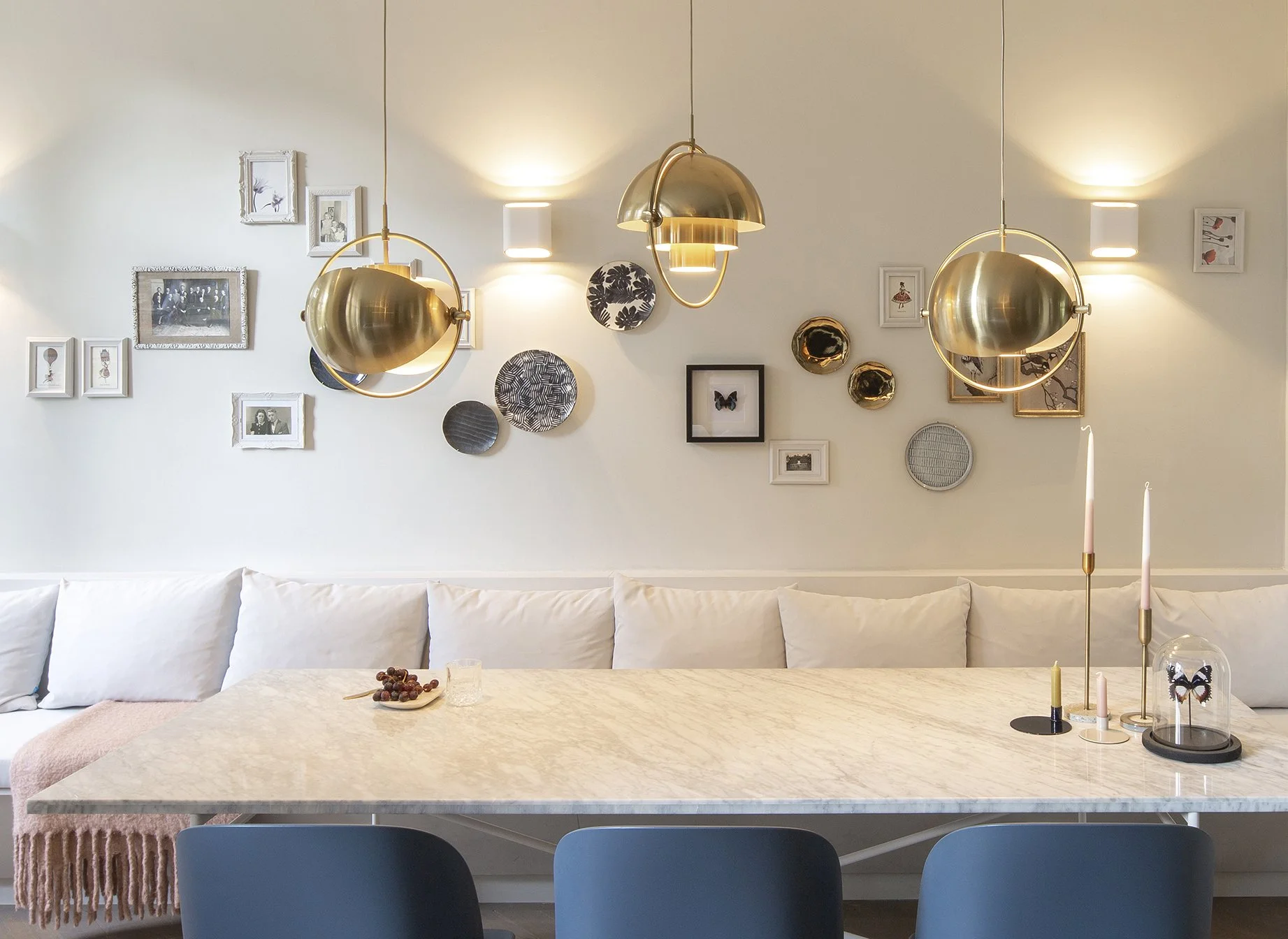 Moderne eetkamer met een witte bank langs de muur, een marmeren tafel met kaarsen en een vaas, en een muur met decoratieve platen, foto's en kunstwerken, verlicht door stijlvolle gouden hanglampen.