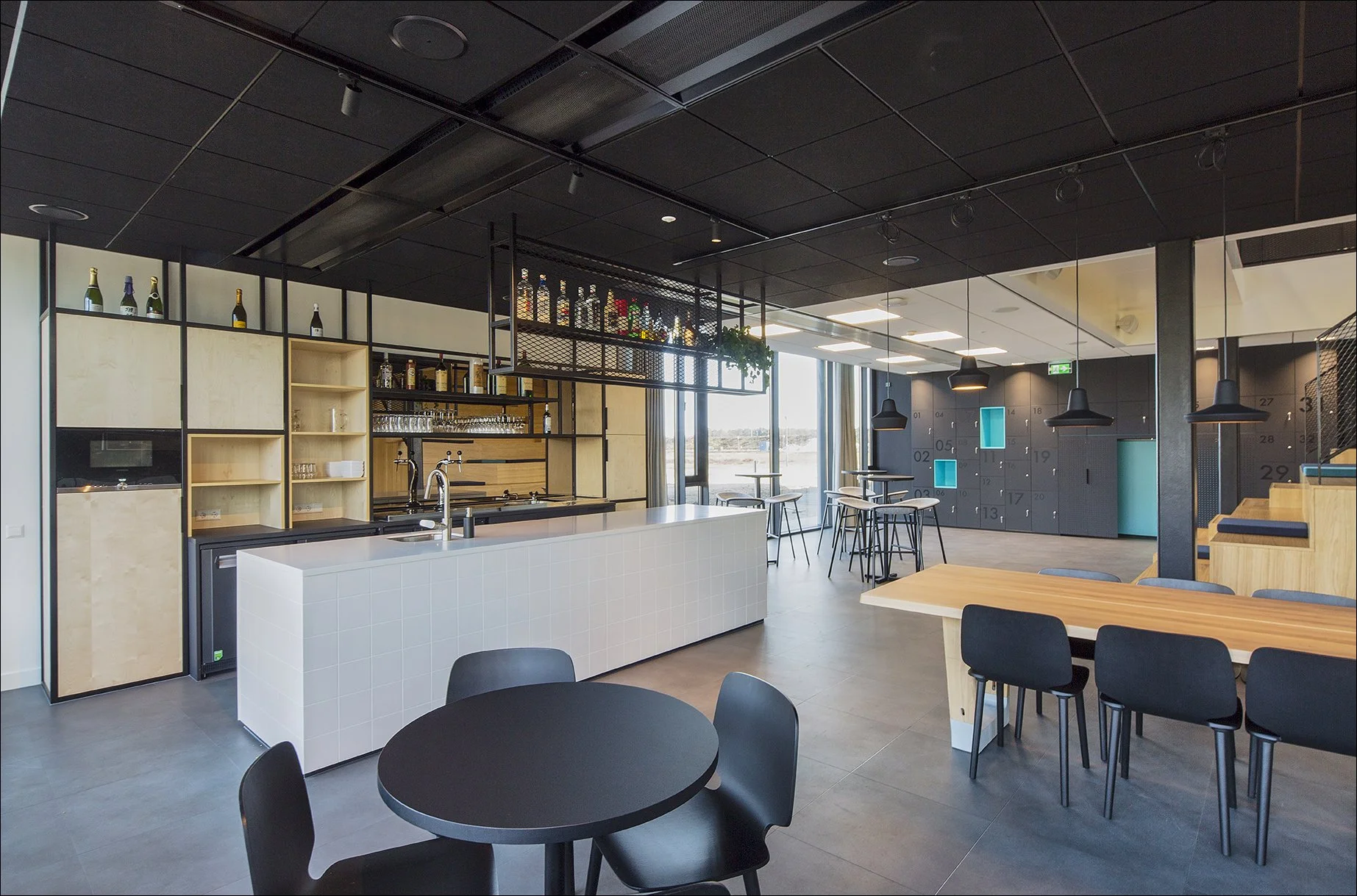 Moderne, lege lounge of café met zwart en hout decor, bar met witte tegels, hoge tafels met krukken, lichtblauwe lockers op de achtergrond, en grote ramen die daglicht binnen laten.