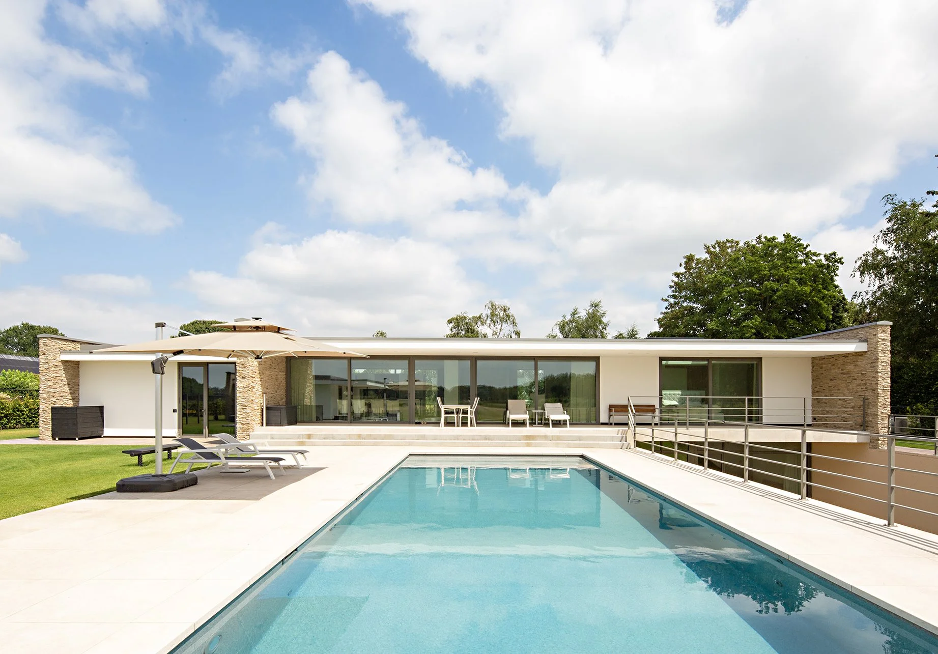 Moderne villa met grote ramen, een zwembad en een terras met ligstoelen en parasol, omgeven door groene bomen onder een blauwe hemel met wolken.